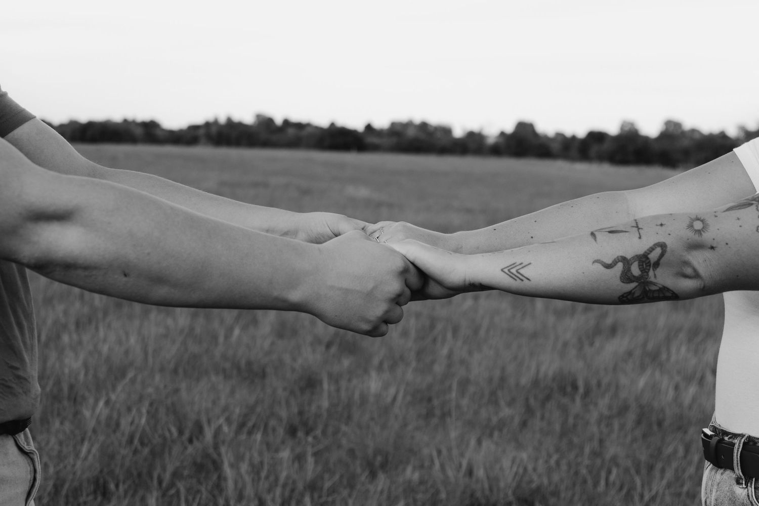 A black and white close-up of tattooed hands holding each other in a field.