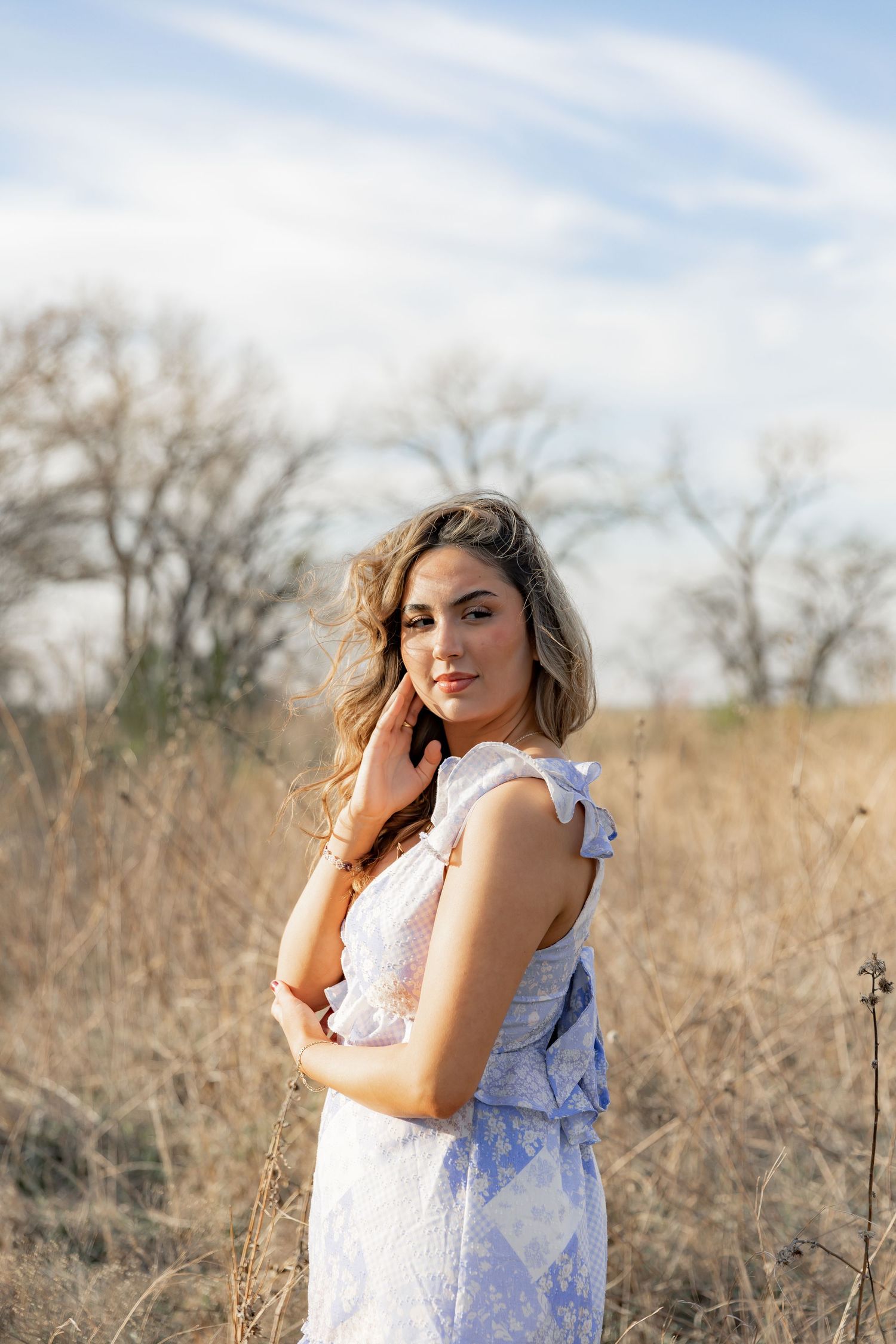 Young woman in blue floral dress posing in golden dry grass field with bare trees and cloudy sky backdrop.