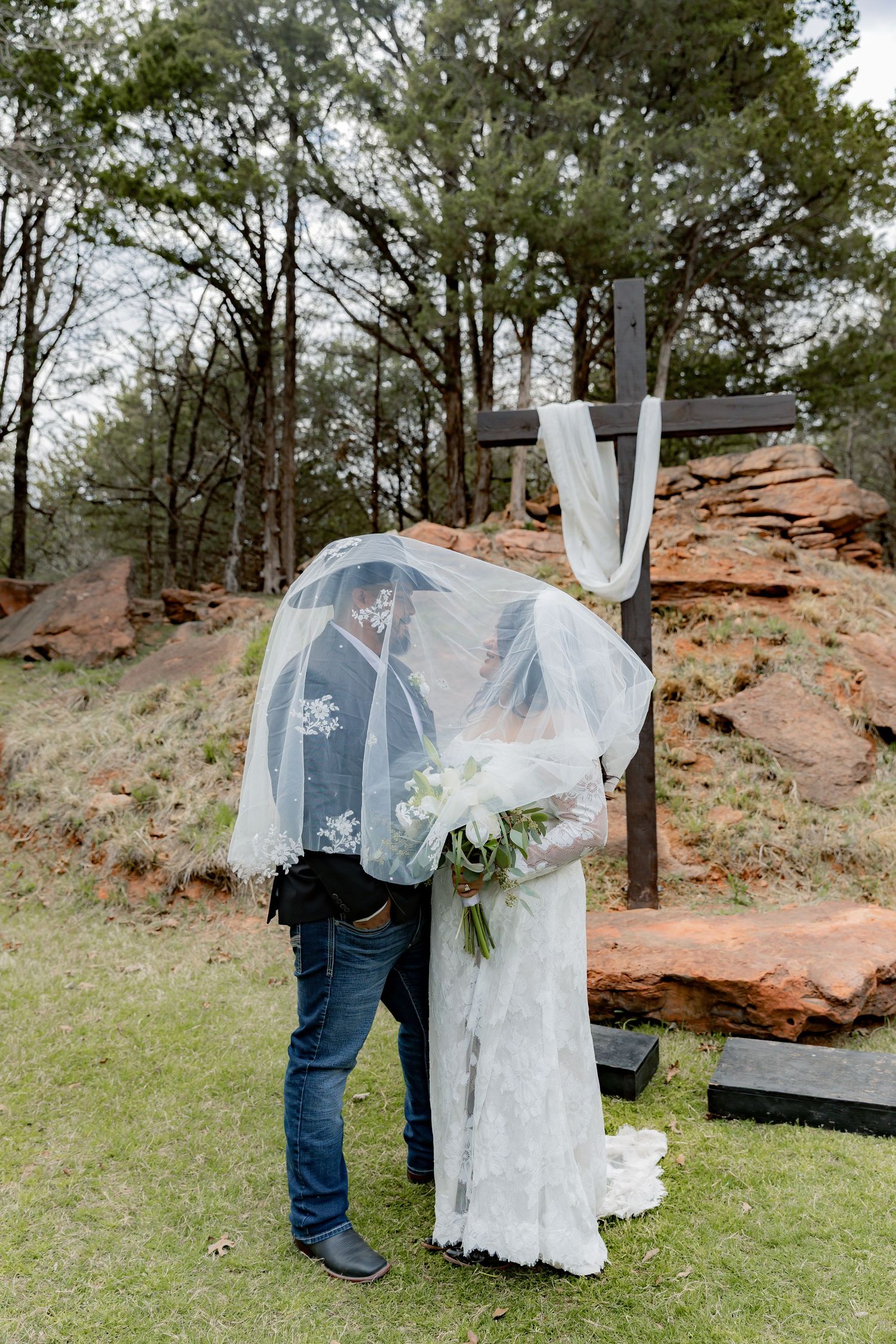 Bride and groom share a kiss under a veil beside a wooden cross at an outdoor wedding ceremony.