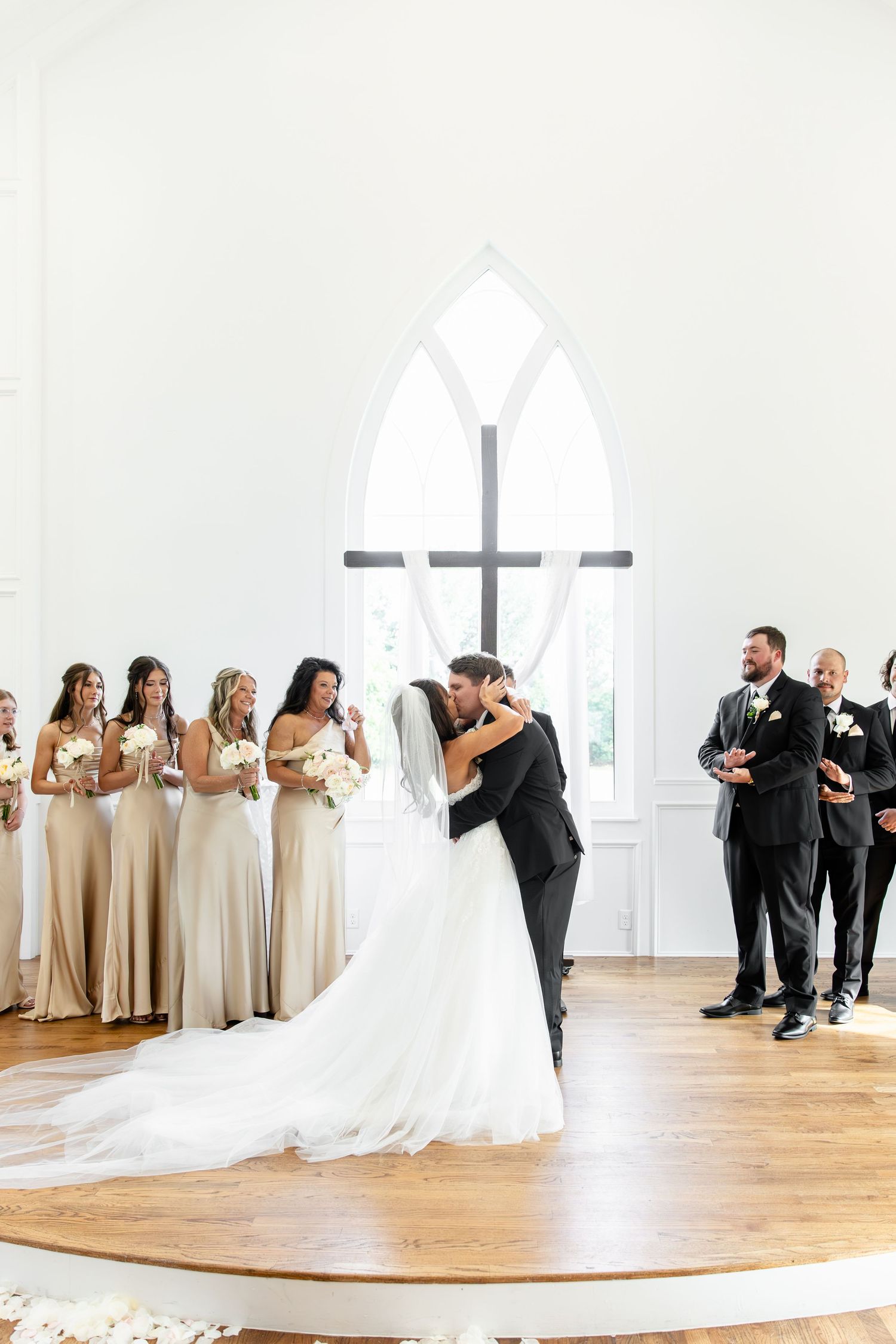 Bride and groom share first kiss at elegant white chapel wedding ceremony with bridesmaids in champagne gowns.