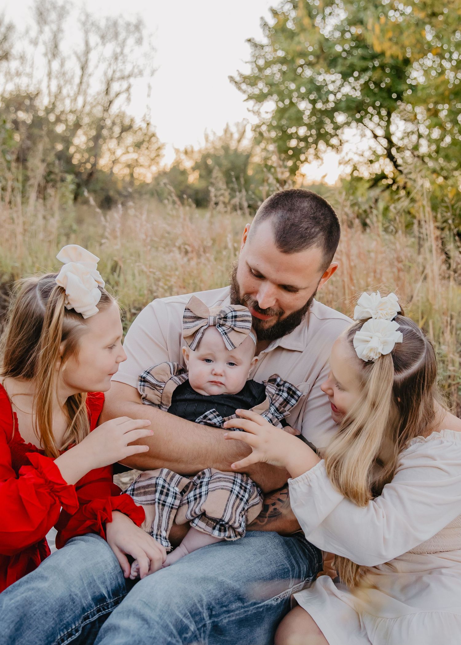 A father sits outdoors in a field holding a baby in plaid while two young girls in bows lean in adoringly.