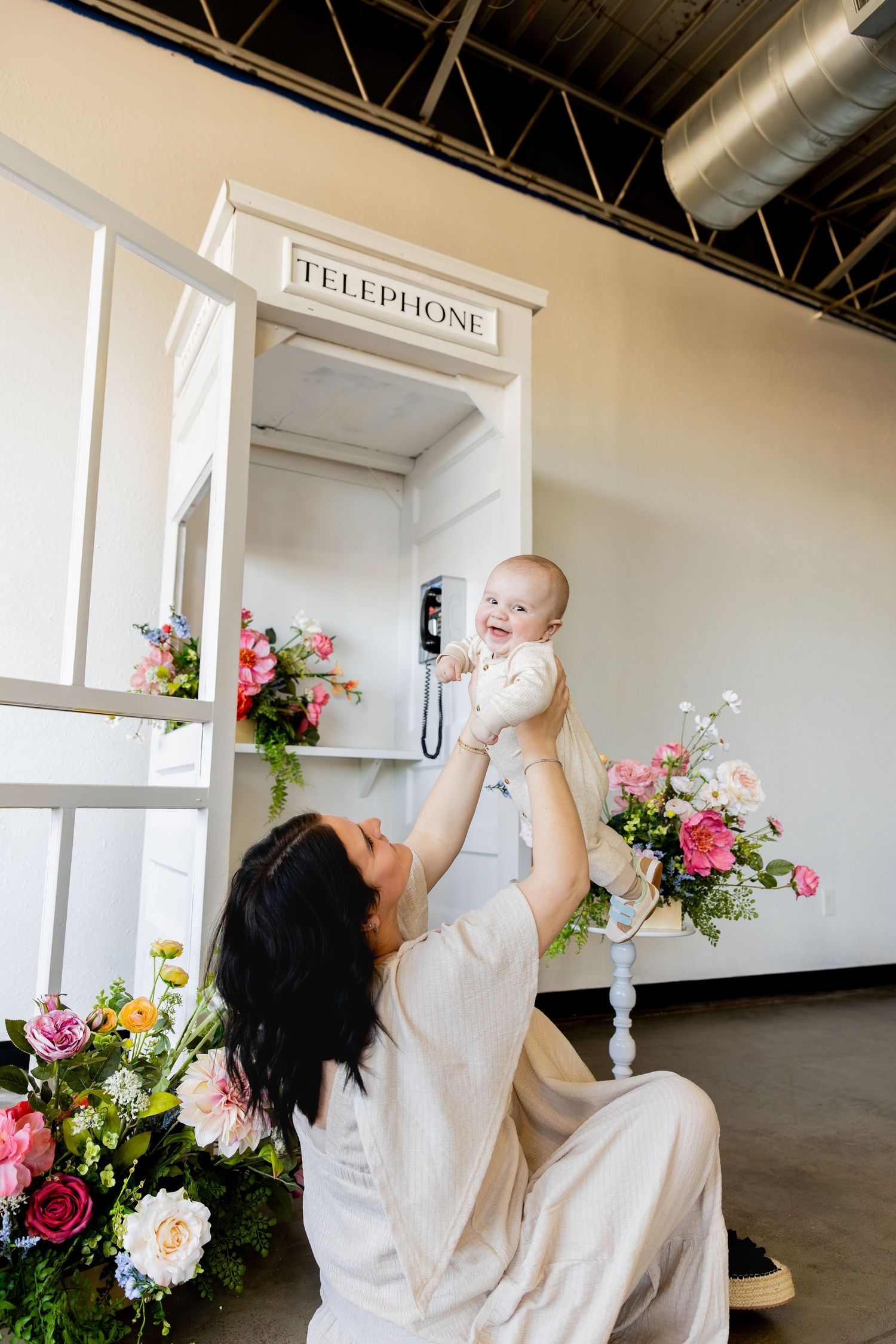 A mother in a white dress holds a baby in white clothing while posing near floral arrangements and a vintage telephone.