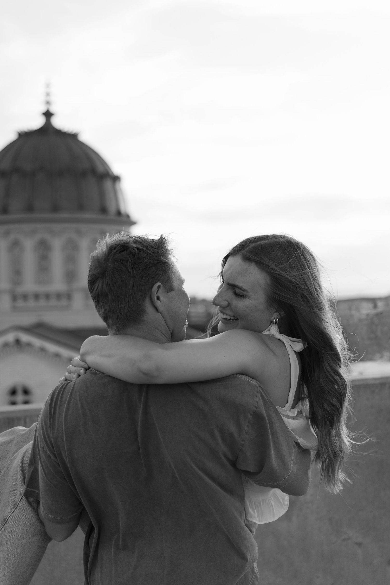 Black and white photo of a couple embracing and laughing with a domed building in the background.