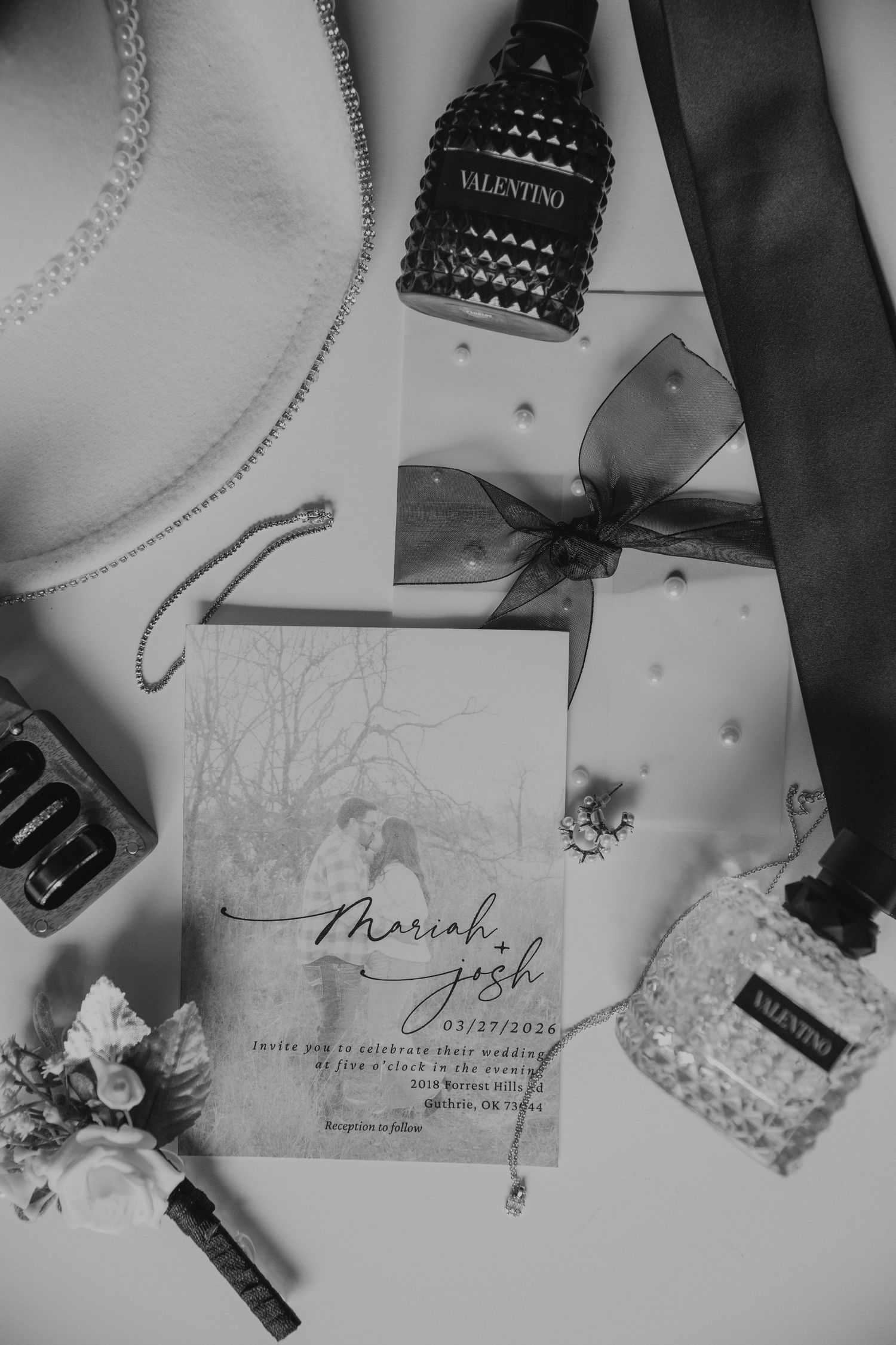 Black and white flat lay of Mariah and Josh wedding invitation suite with Valentino perfume, tie, and floral boutonniere.
