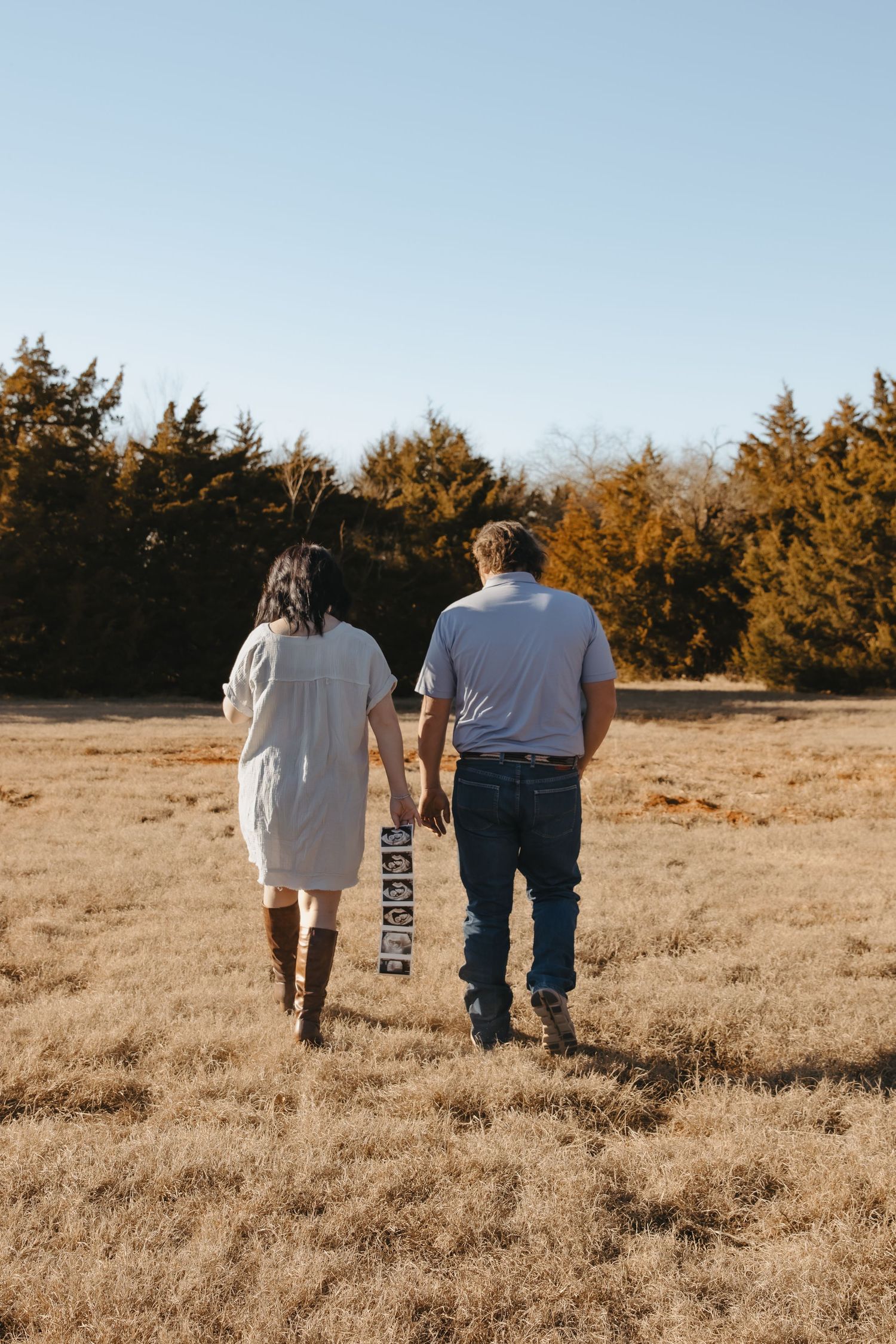 Couple holding hands walking through golden field toward evergreen trees, woman in white dress and boots, man in jeans.