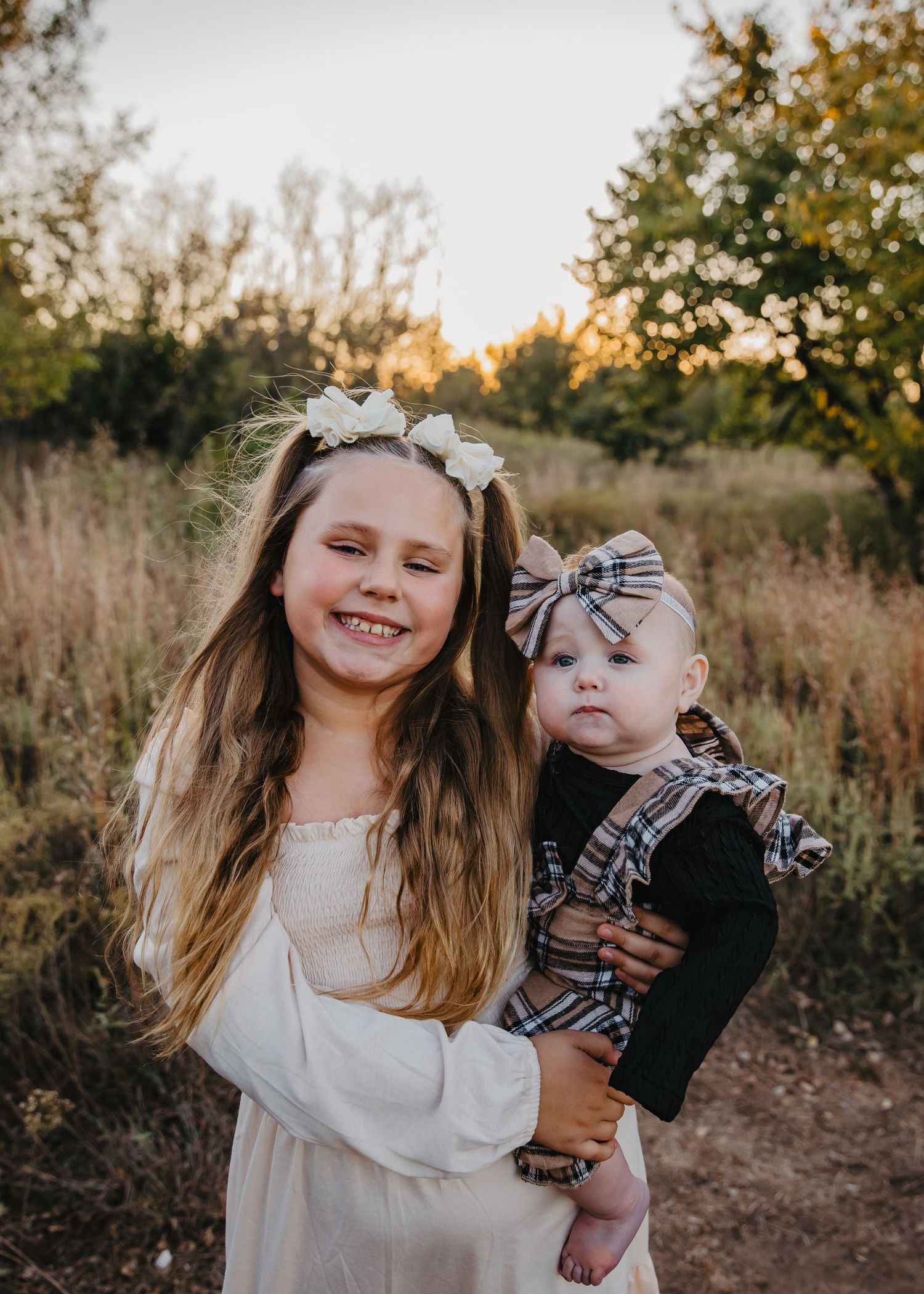 A smiling girl in white holds her baby sister in plaid outdoors at sunset.