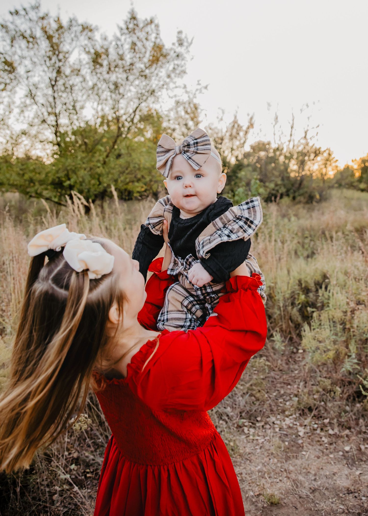 A mother in a red dress lifts her plaid-dressed baby girl outdoors at golden hour.