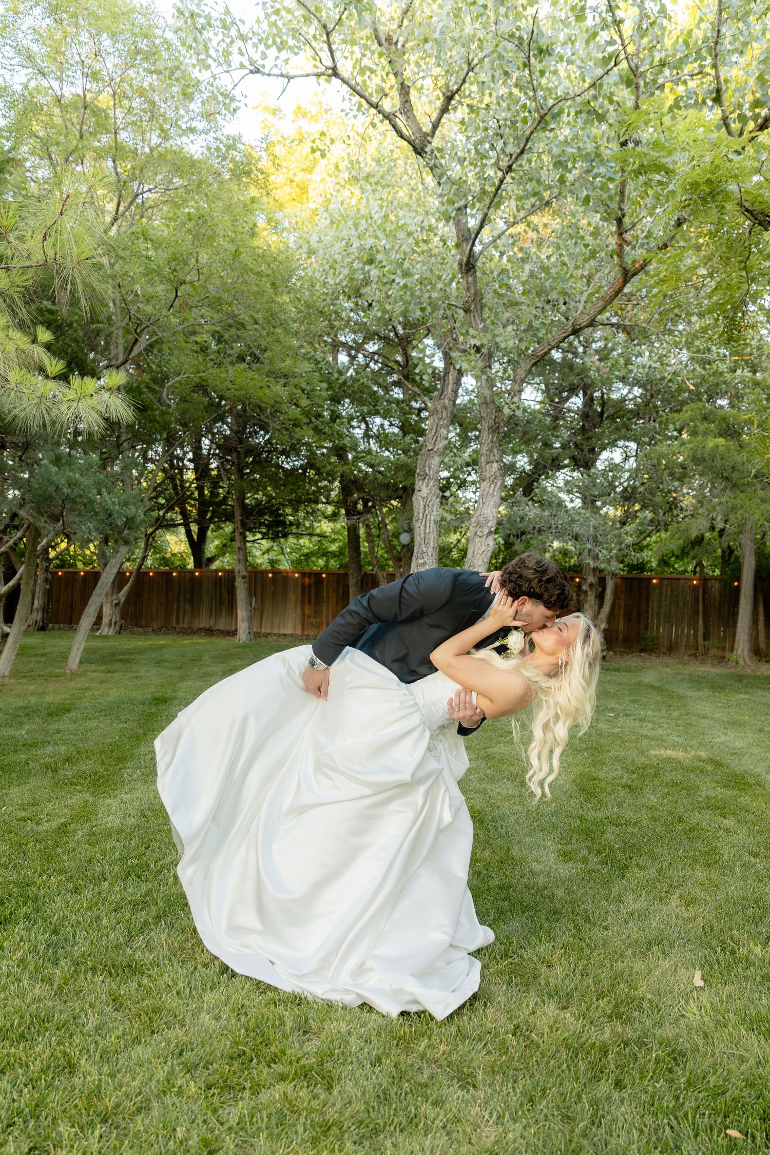 Groom dips bride in white gown for a romantic kiss on green lawn surrounded by lush trees at outdoor wedding.