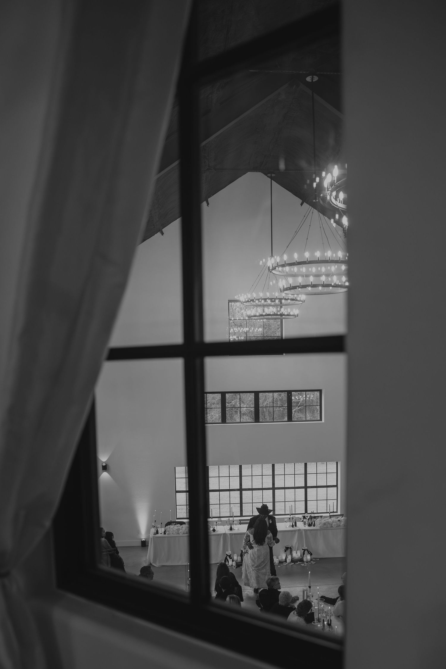 Black and white photo of a wedding reception viewed through a window, featuring elegant chandeliers and dancing couple.