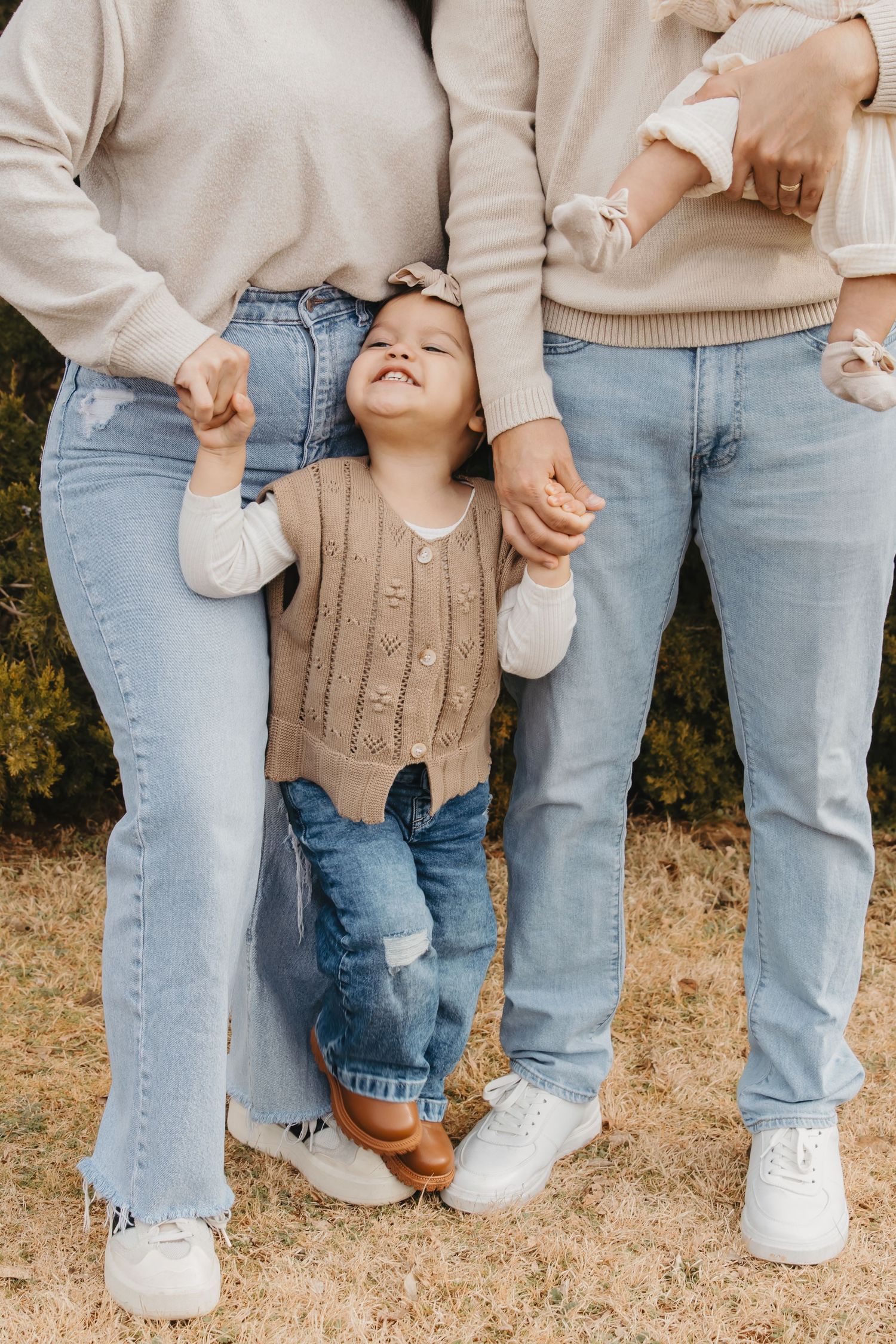 Small child in brown sweater holding hands with parents, wearing matching denim and neutral tones.