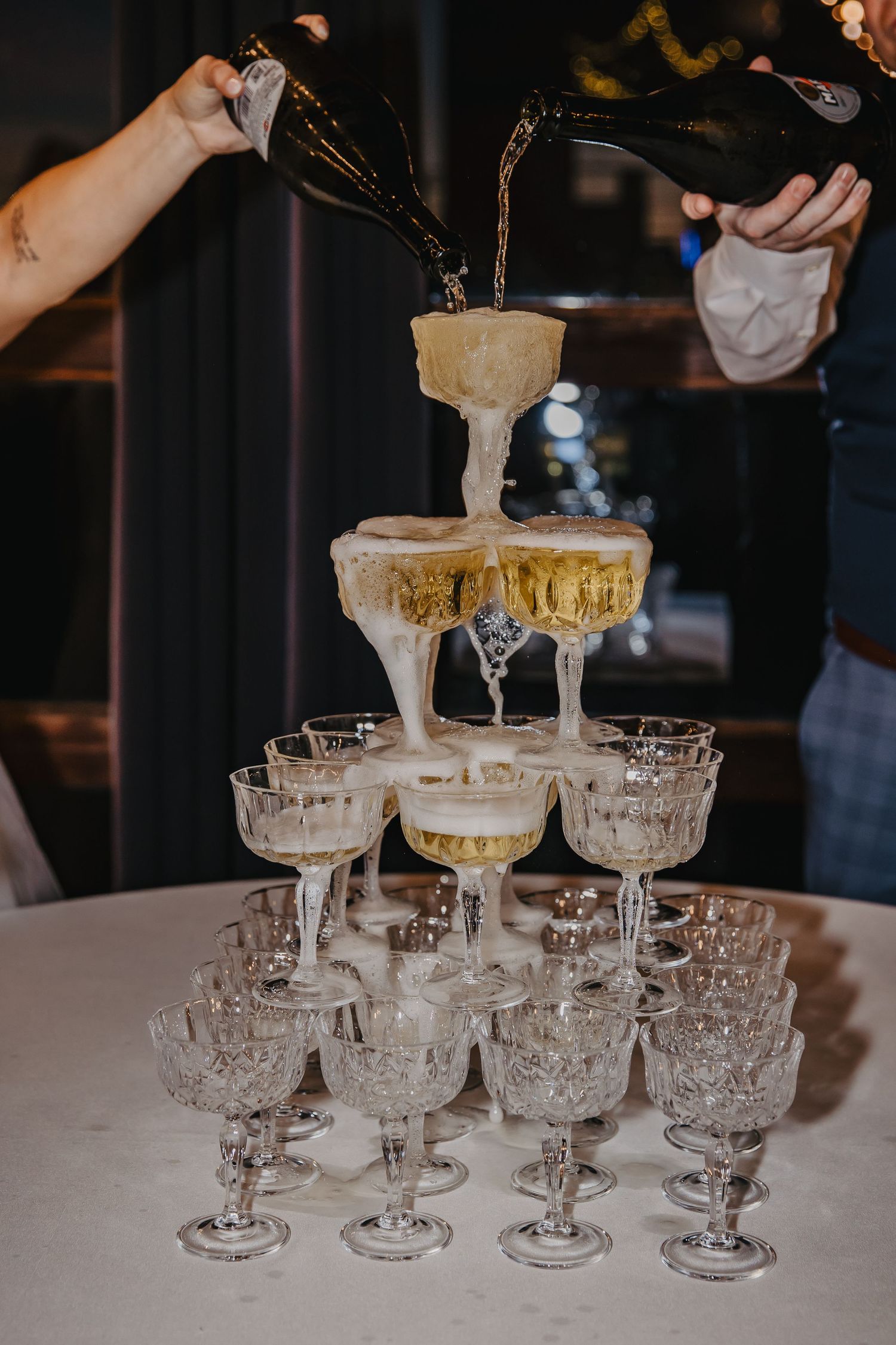 Champagne tower with crystal glasses being filled by two people pouring bottles at an elegant event.