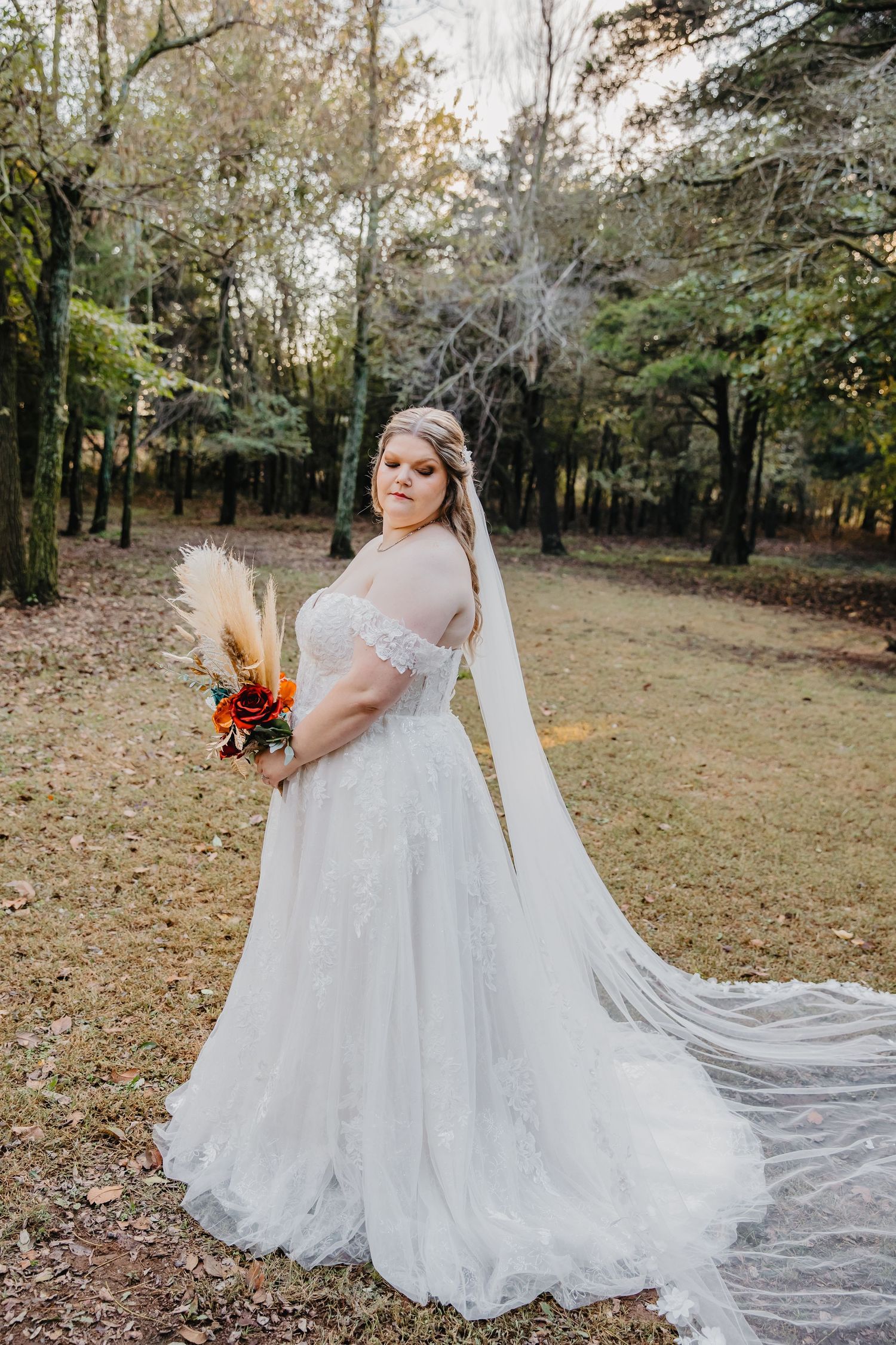 Bride in white lace gown with long veil holds boho bouquet in autumn forest setting.