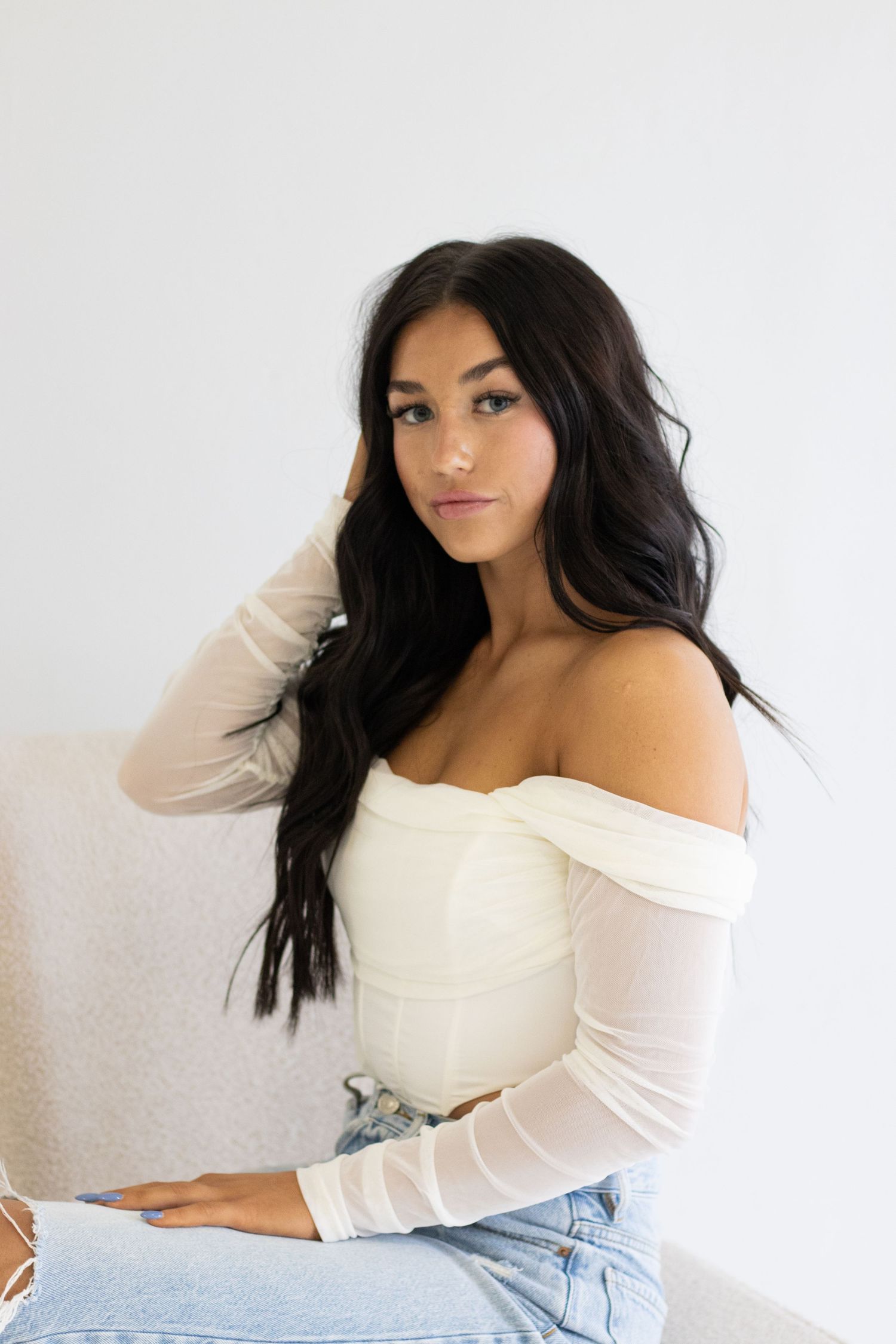 Portrait featuring off-shoulder white top and long dark hair against minimal background.