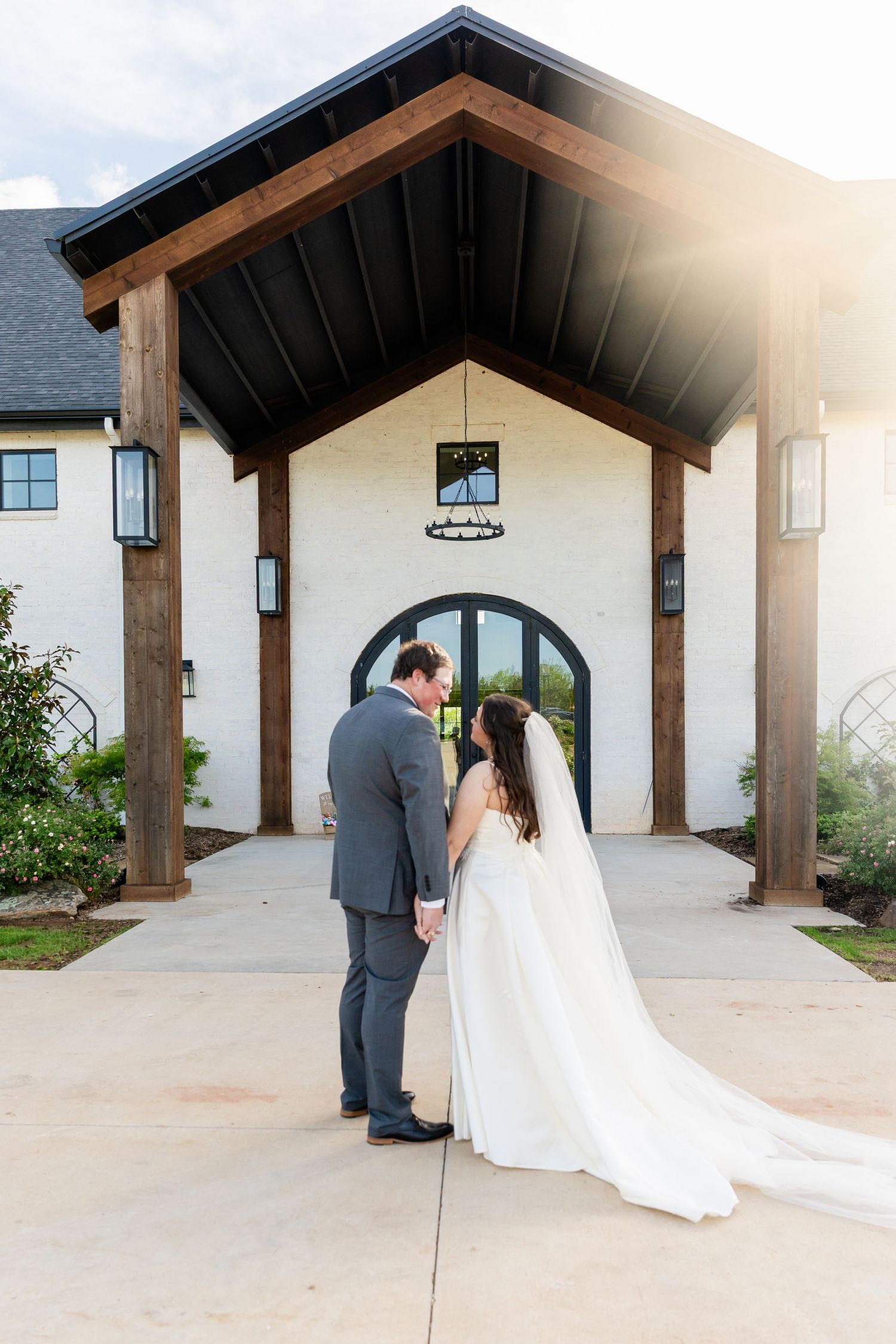Bride and groom hold hands facing a white wedding venue with wooden beams and arched doors in warm sunlight.