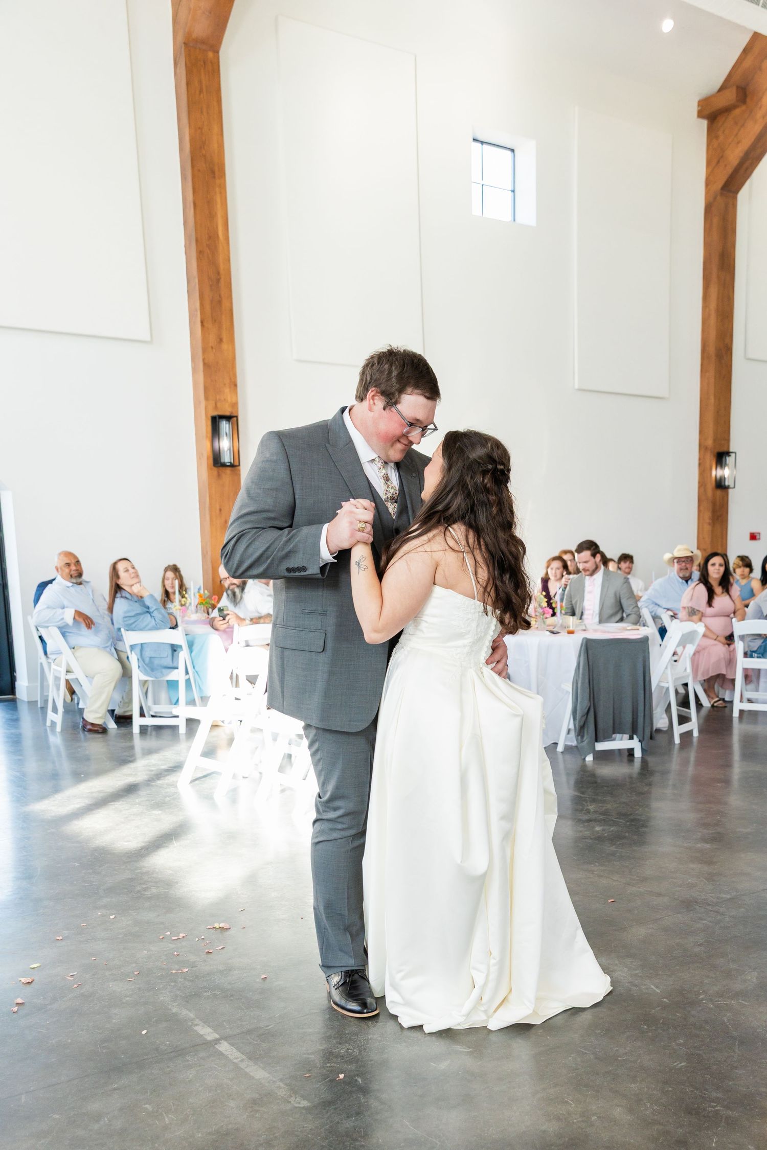 Bride and groom share their first dance in a bright white barn wedding venue with wooden beams and seated guests.