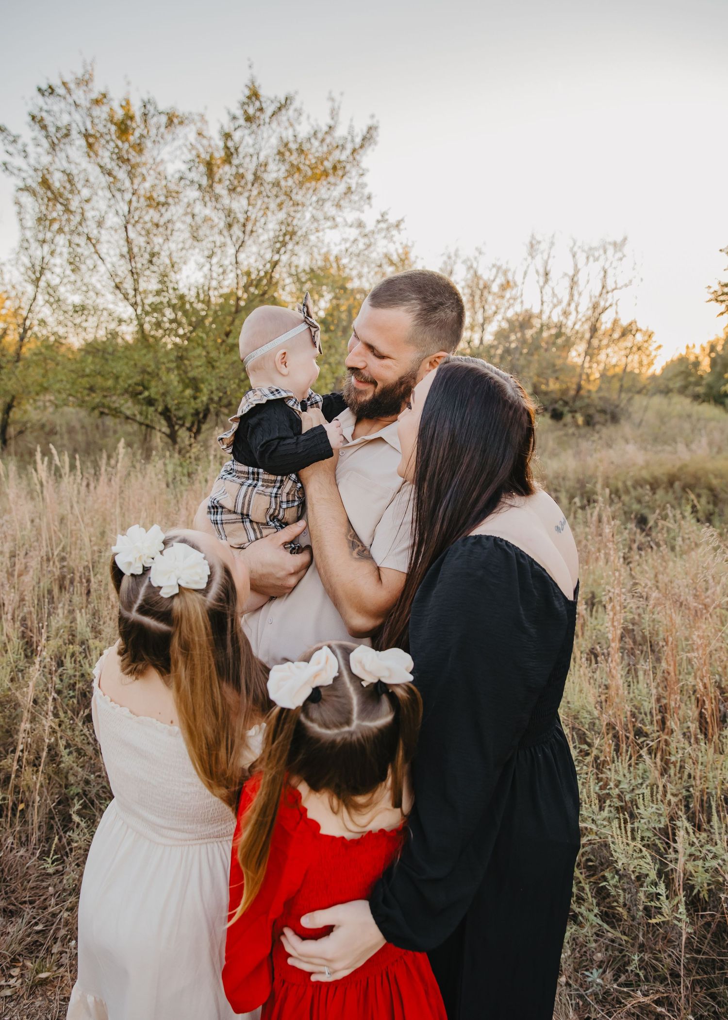 Family portrait at golden hour in a field, parents holding baby and hugging two young girls in dresses.