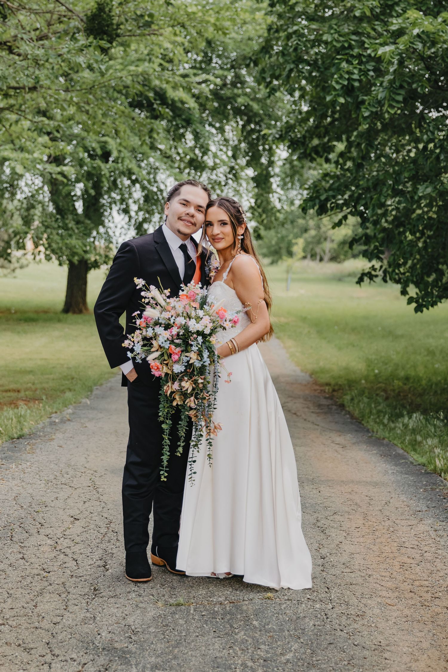A wedding couple poses on a dirt path surrounded by lush green trees holding a cascading pink and white floral bouquet.