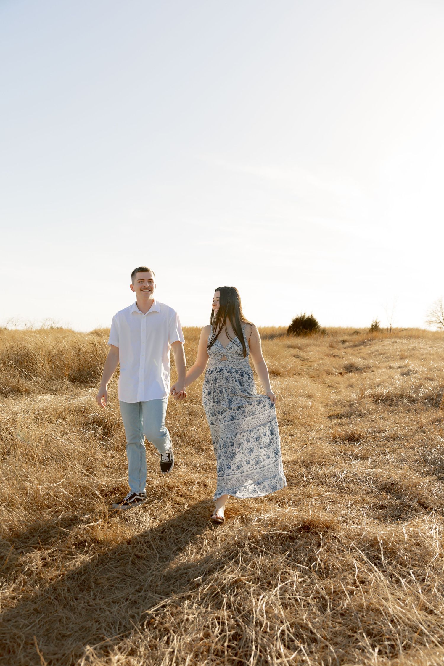 Couple holding hands walking through golden dry grass field, man in white shirt, woman in floral maxi dress, sunny day.