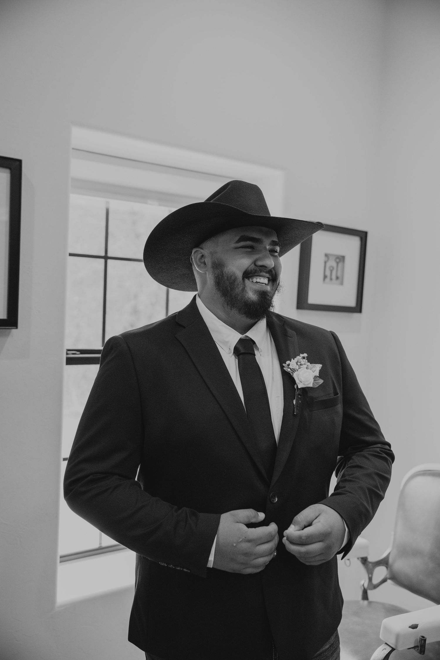 Smiling groom in black suit, cowboy hat, and boutonniere buttoning jacket in black and white wedding photo.