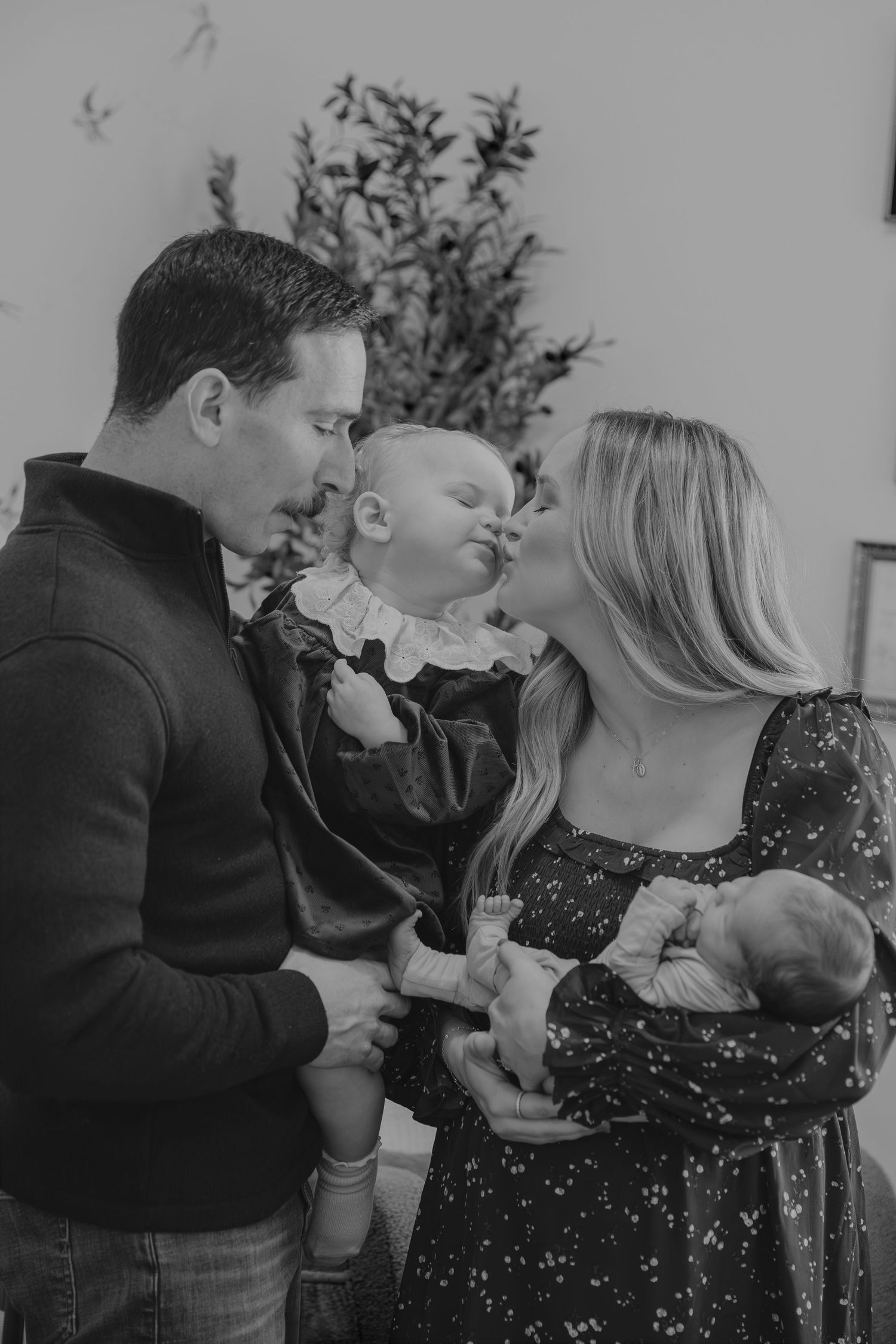 Black and white family photo of parents kissing toddler while mother holds newborn baby indoors.