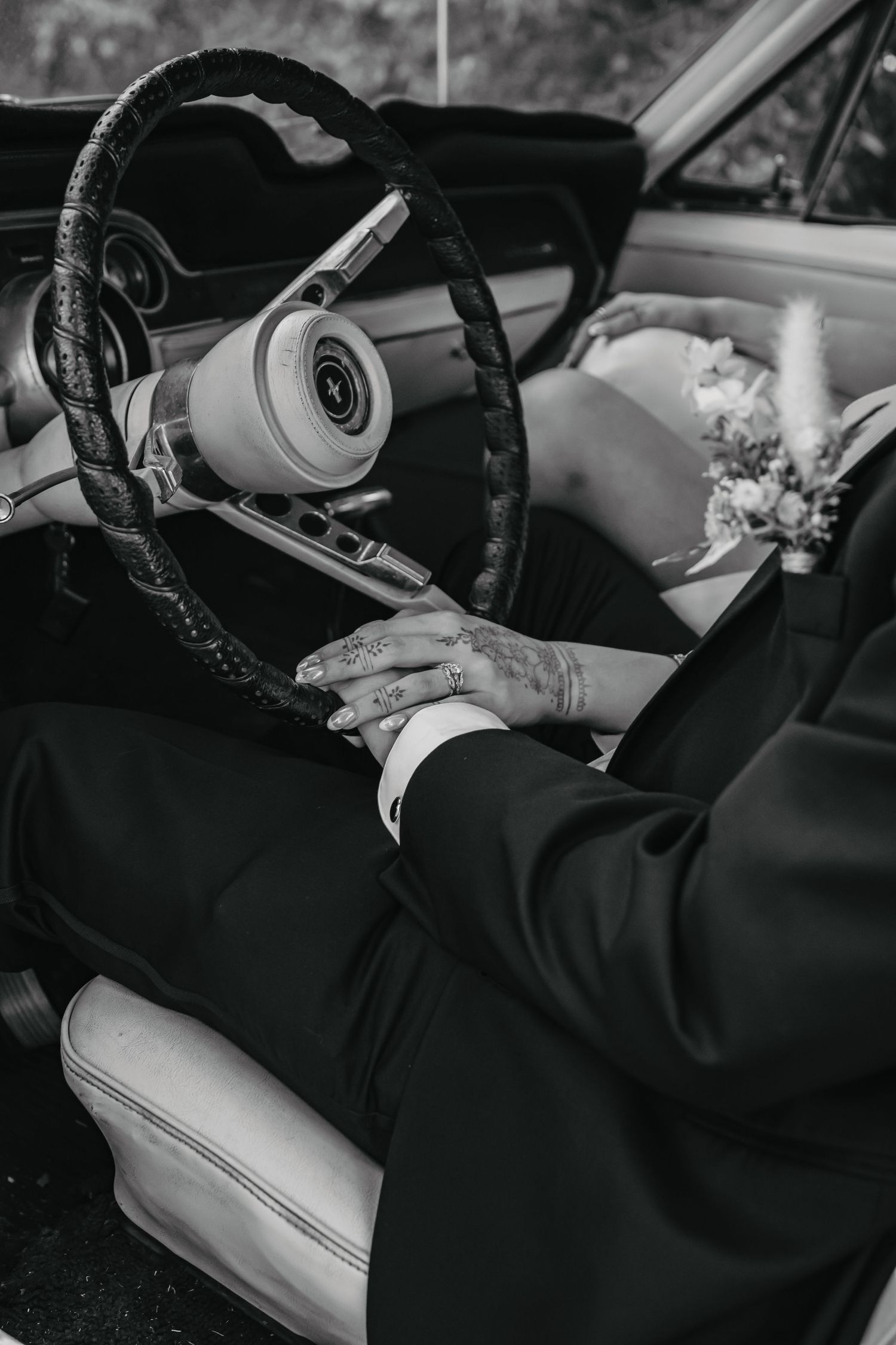 A black and white close-up of hands on a vintage car steering wheel with white leather interior.