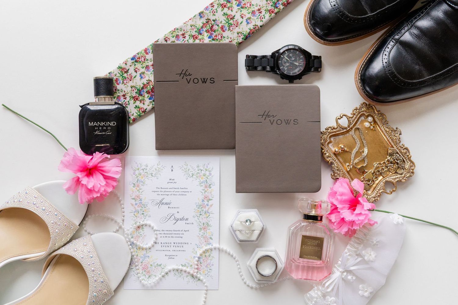 Wedding flat lay with vow books, invitations, perfume, watch, shoes, rings, and pink flowers on white background.