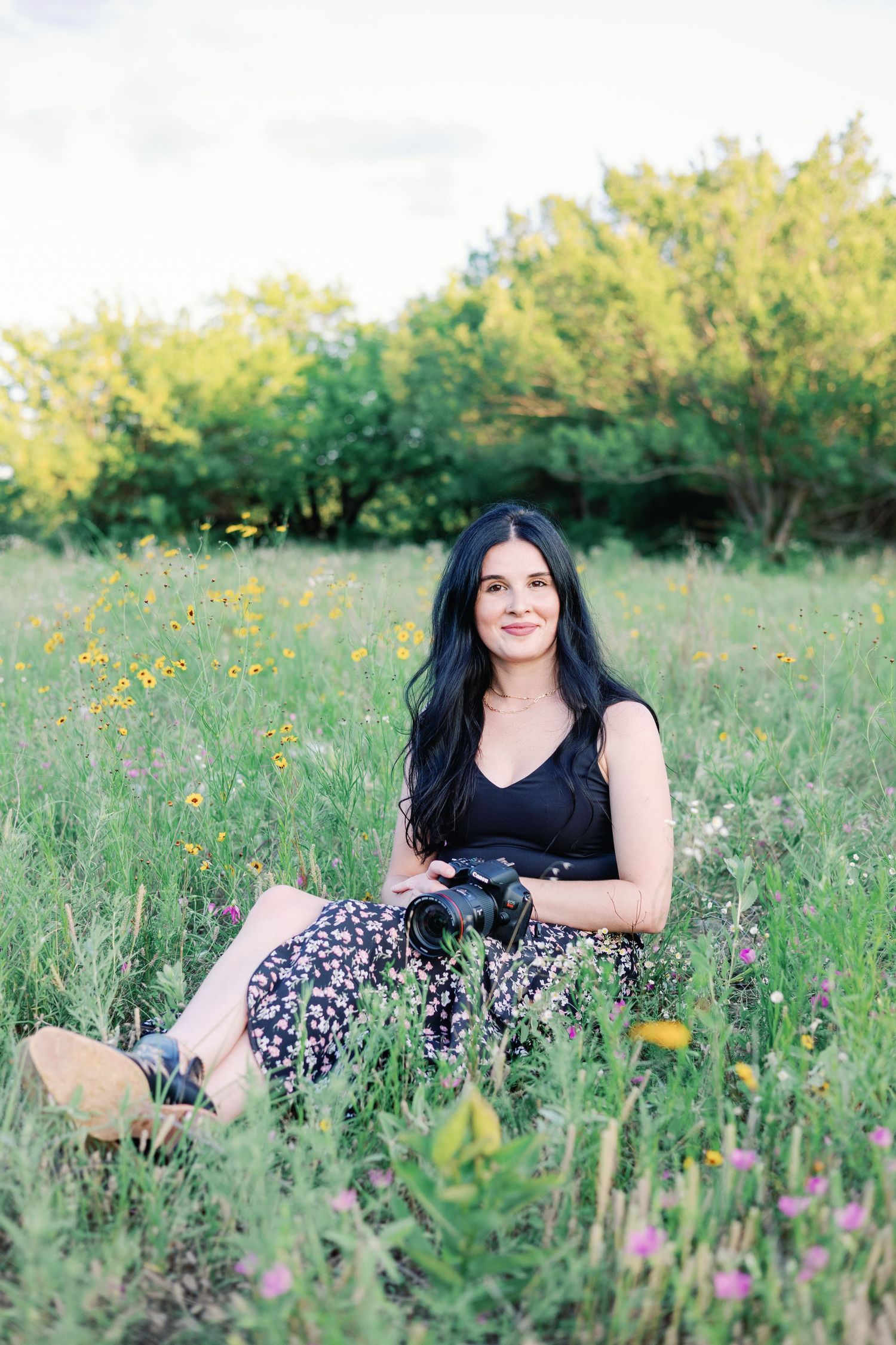 A photographer in a black top and floral skirt sits among wildflowers holding a camera.