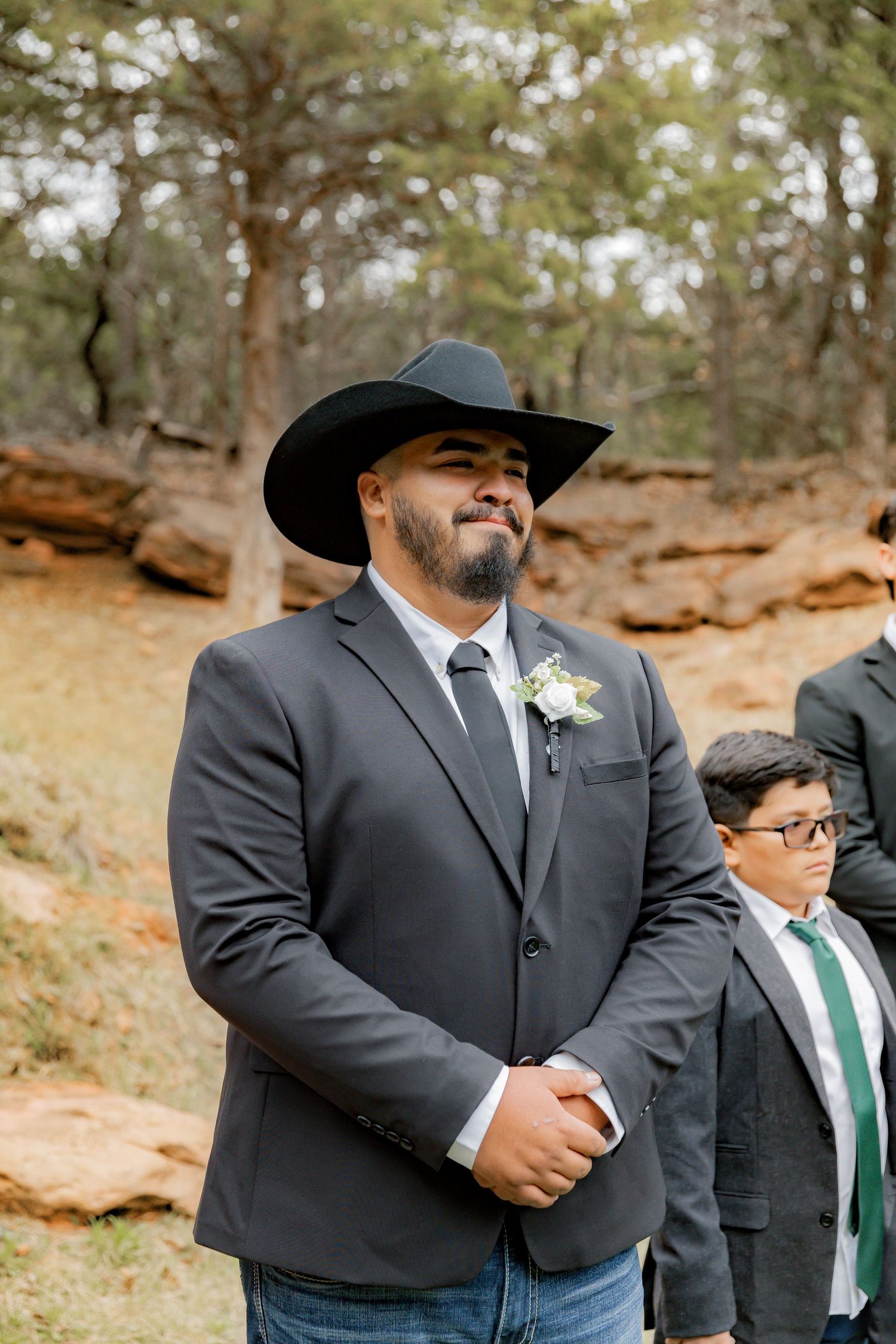 Groom in black cowboy hat and gray suit with boutonniere smiling at outdoor rustic wedding ceremony.