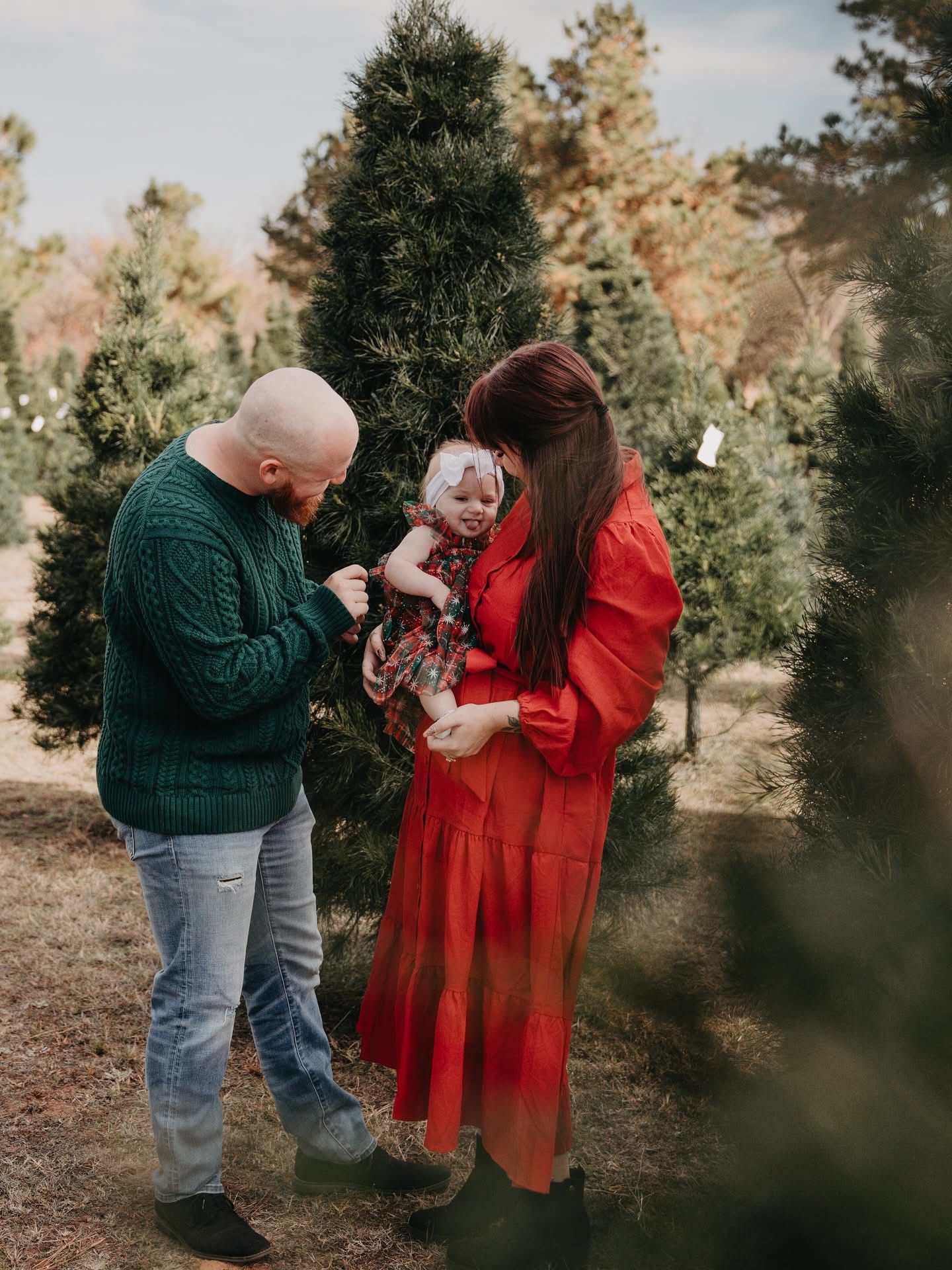 Family with baby at Christmas tree farm, parents in red and green holding infant in floral dress among evergreen trees.