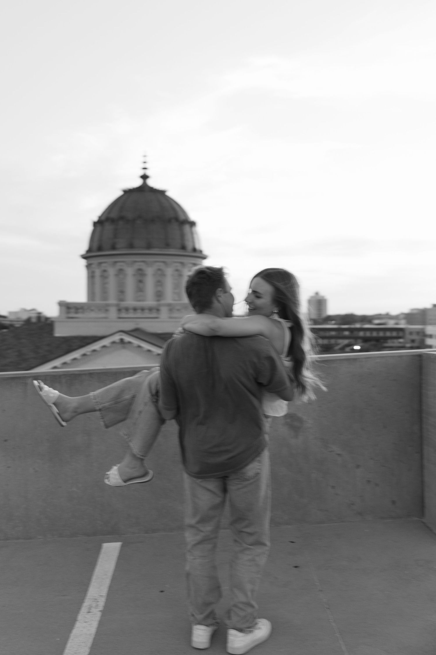 A man romantically lifts a woman on a rooftop with a domed building in the background in this black and white photo.