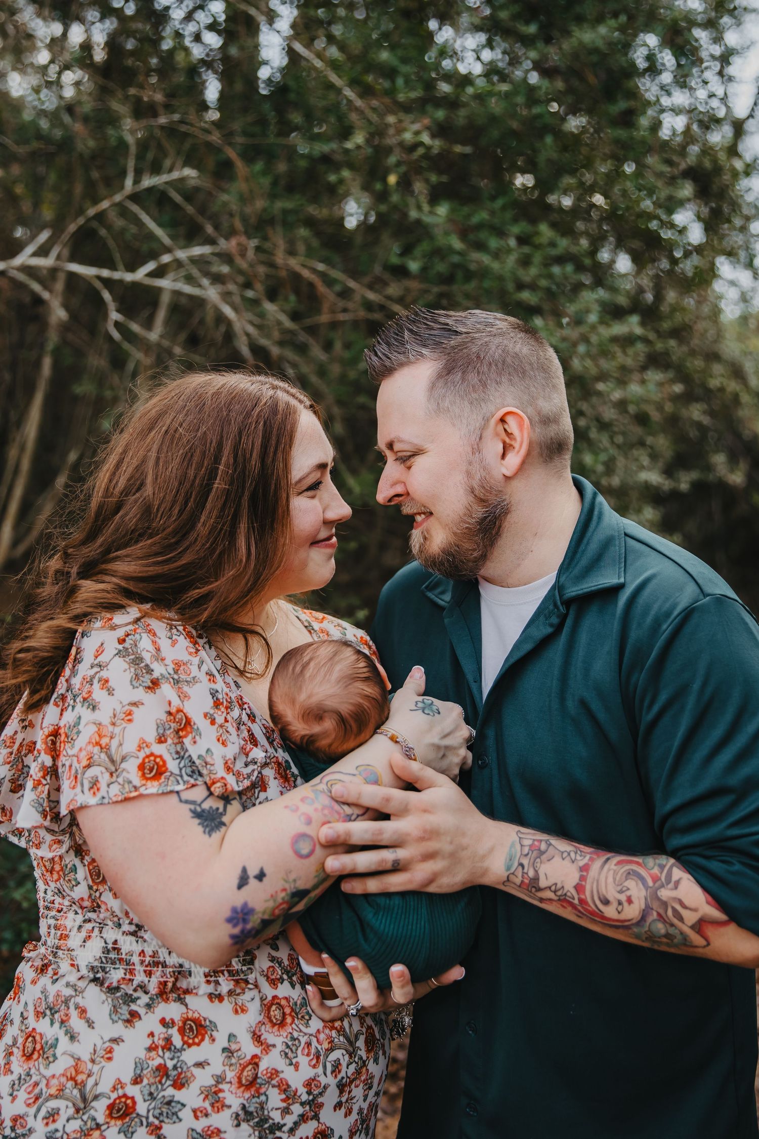 Couple with tattoos smiling at each other while holding their newborn baby outdoors near green trees.