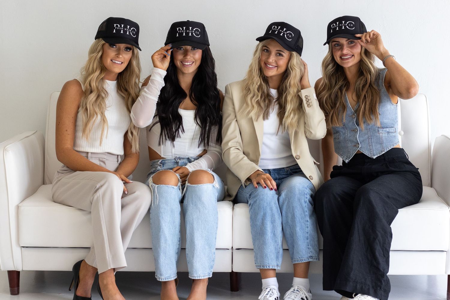 Four women wearing matching black caps and denim sitting on white couch.