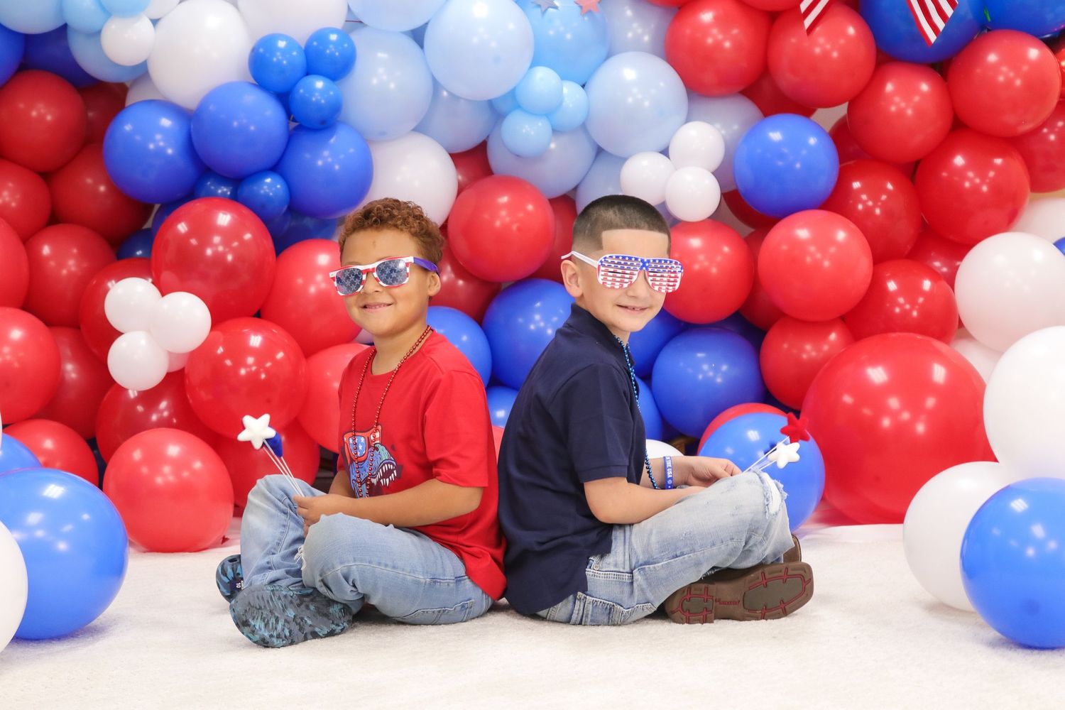 Two children sitting back-to-back against a backdrop of red, white and blue balloons for a patriotic themed photo.