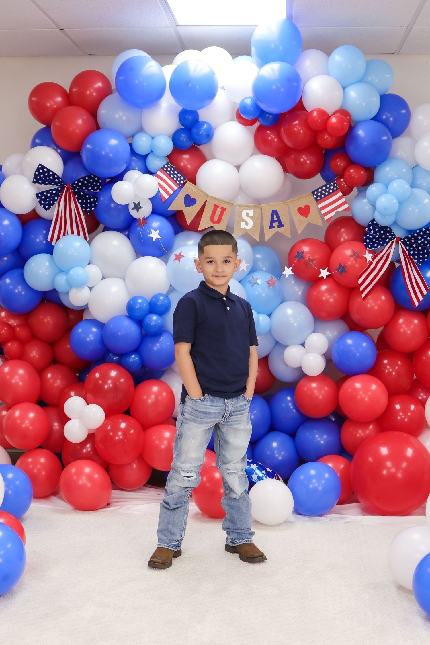 Standing child against red white and blue balloon wall with USA banner.