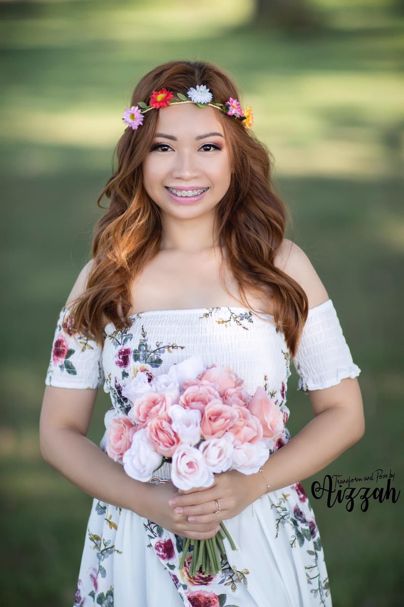 Woman in white floral dress holding pink roses with flower crown in garden setting.