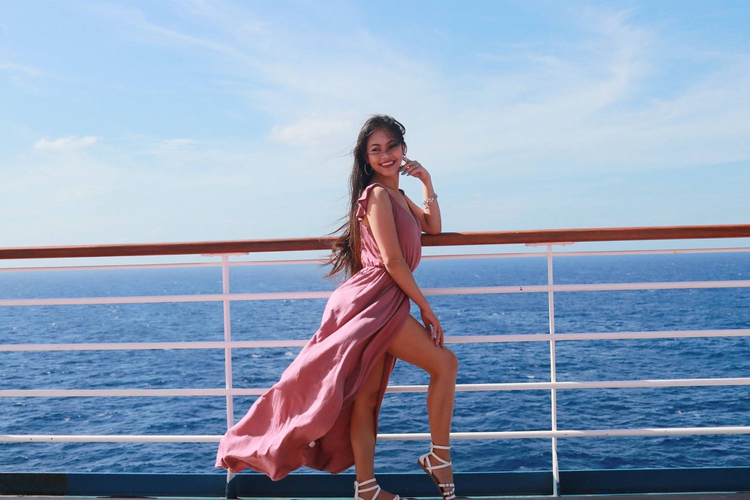 A flowing pink dress catches the sea breeze on a cruise ship deck with blue ocean views.