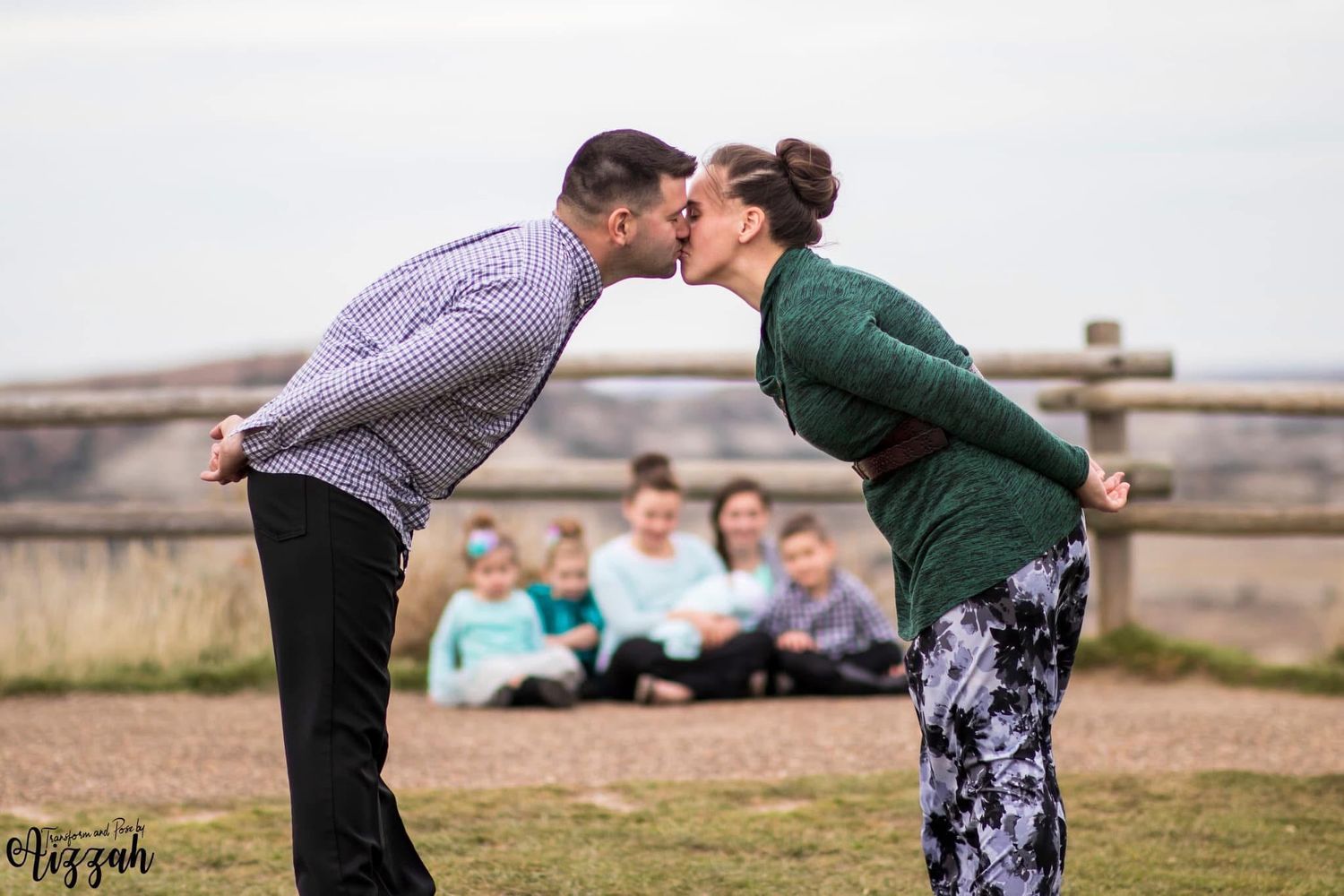 Playful couple leaning in for kiss with family sitting in background outdoors.