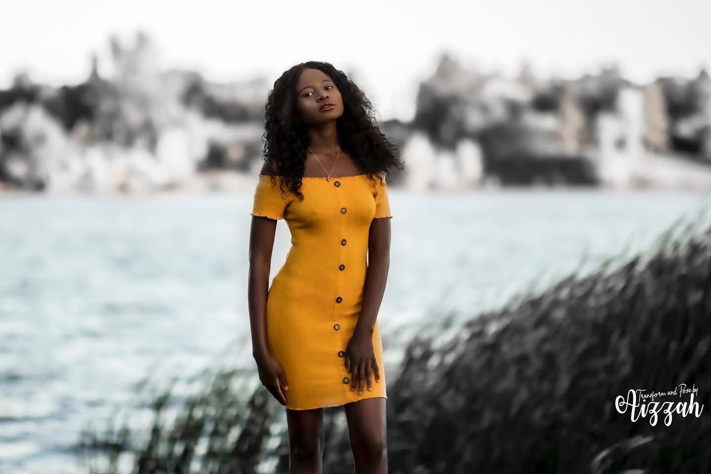 Portrait of woman in yellow button-down dress standing by waterfront with urban backdrop.