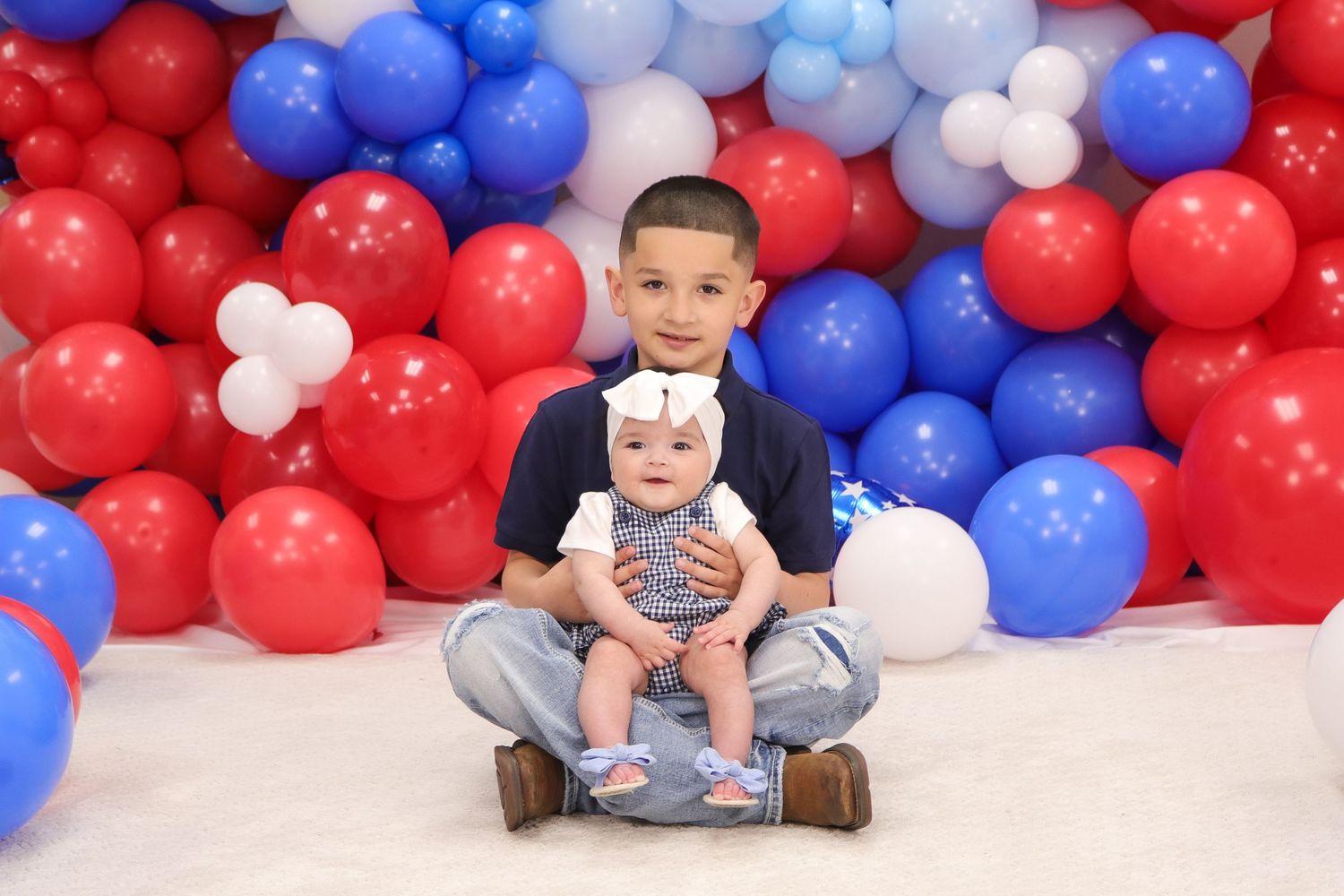 Child sitting with baby doll against patriotic balloon backdrop.