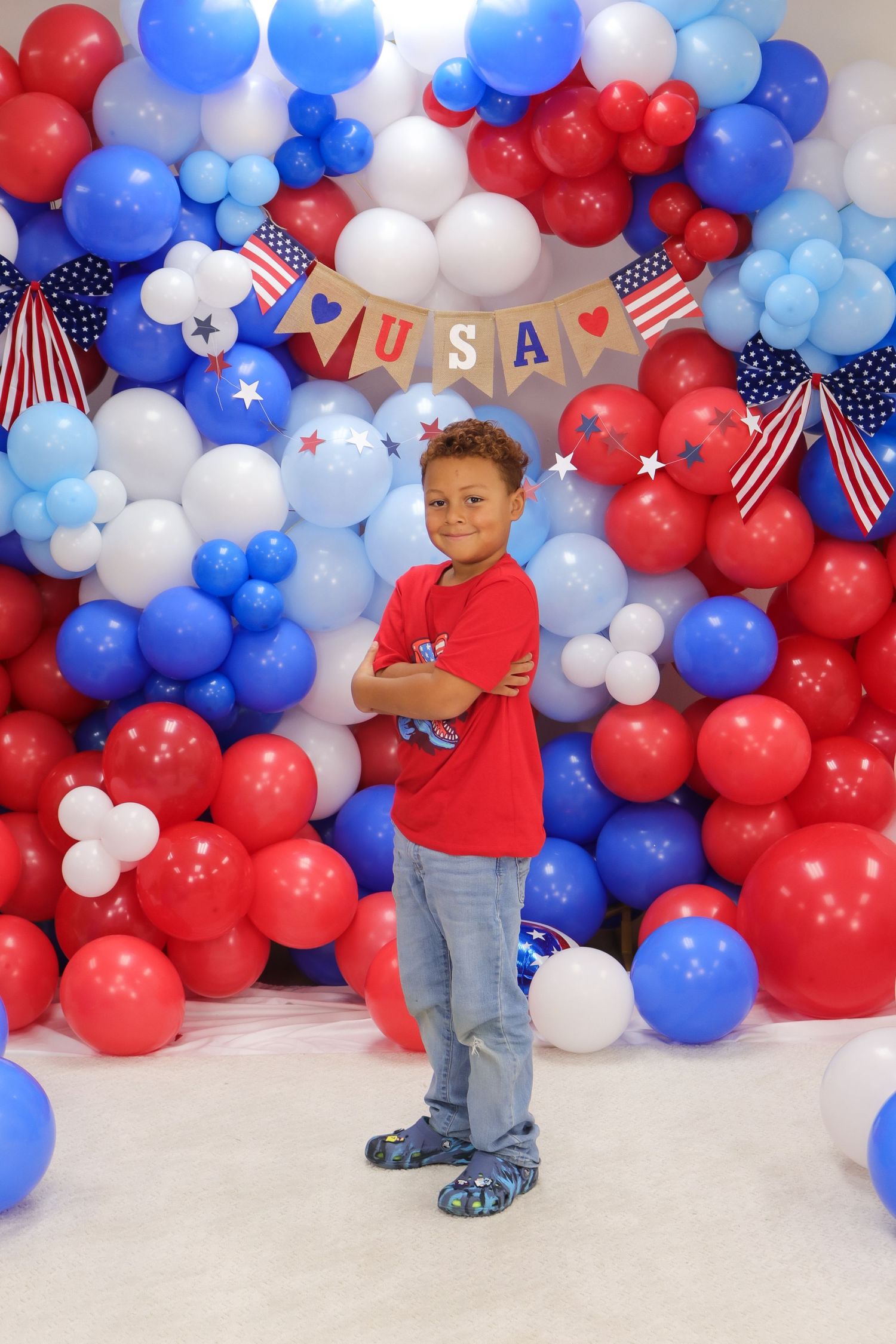 Child in red shirt poses confidently against patriotic balloon backdrop.