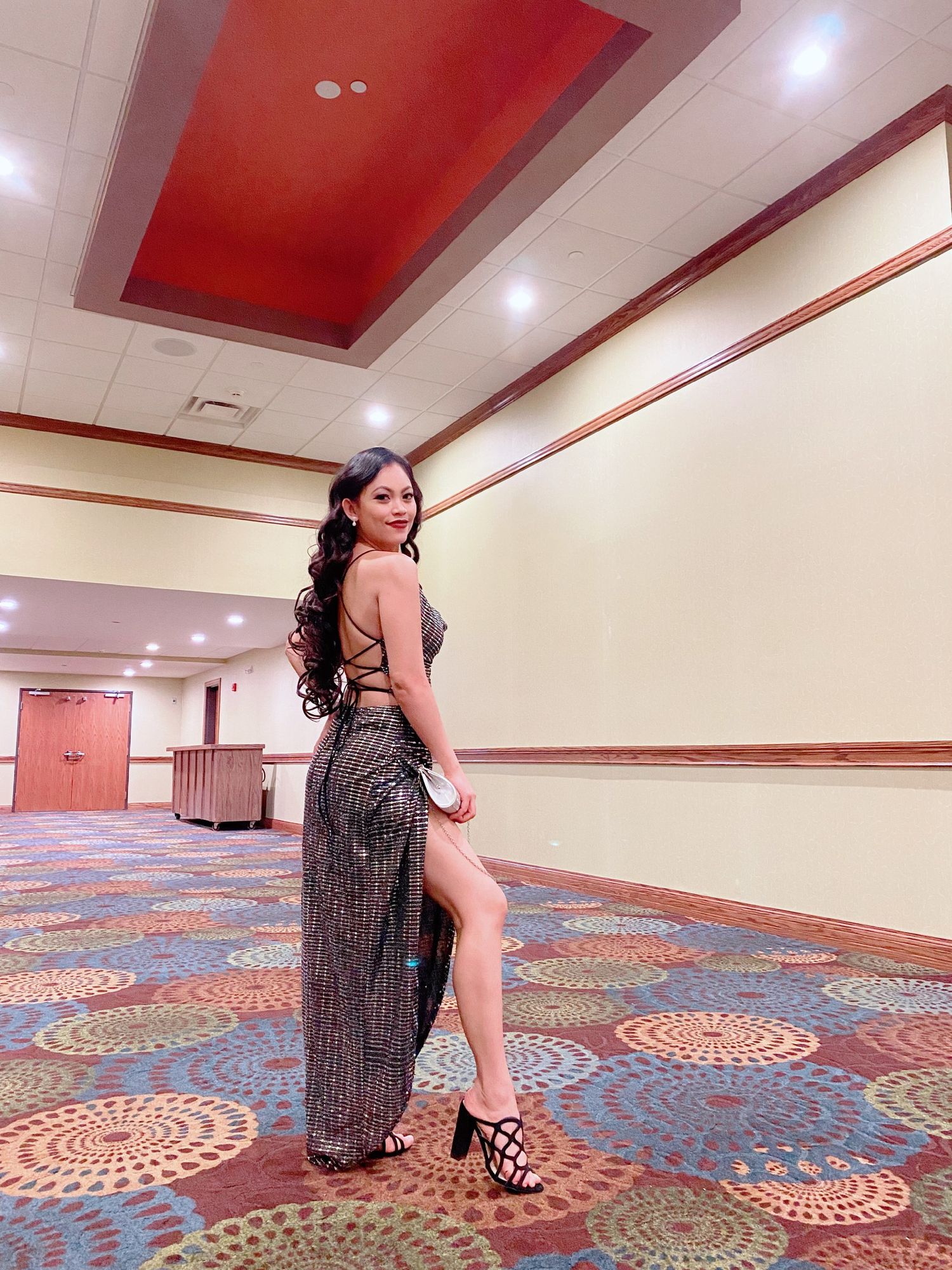 Elegant evening dress pose in a hotel ballroom with patterned carpet and red ceiling accents.