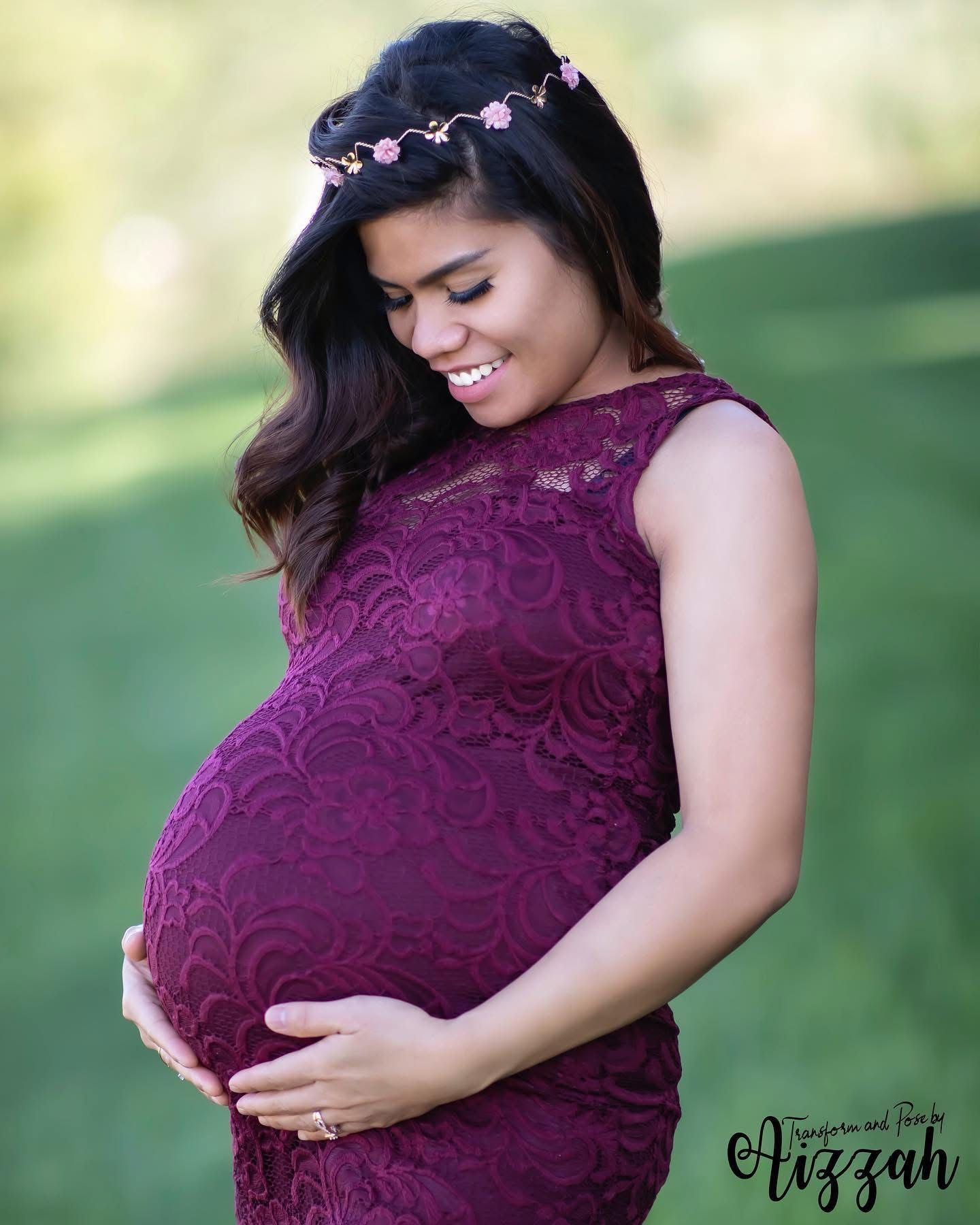Expectant mother in purple lace dress smiles while cradling baby bump outdoors.