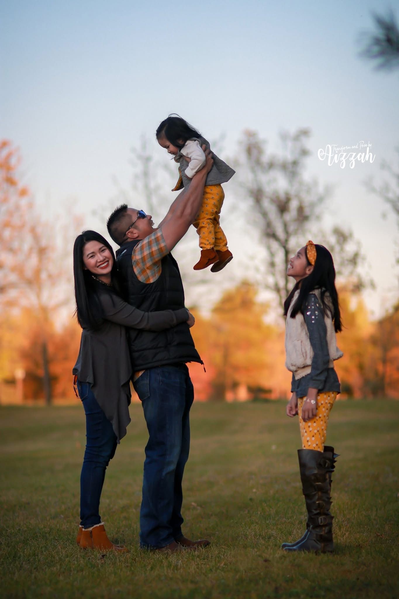 Family playing outdoors at sunset with parent lifting child in air while others watch.