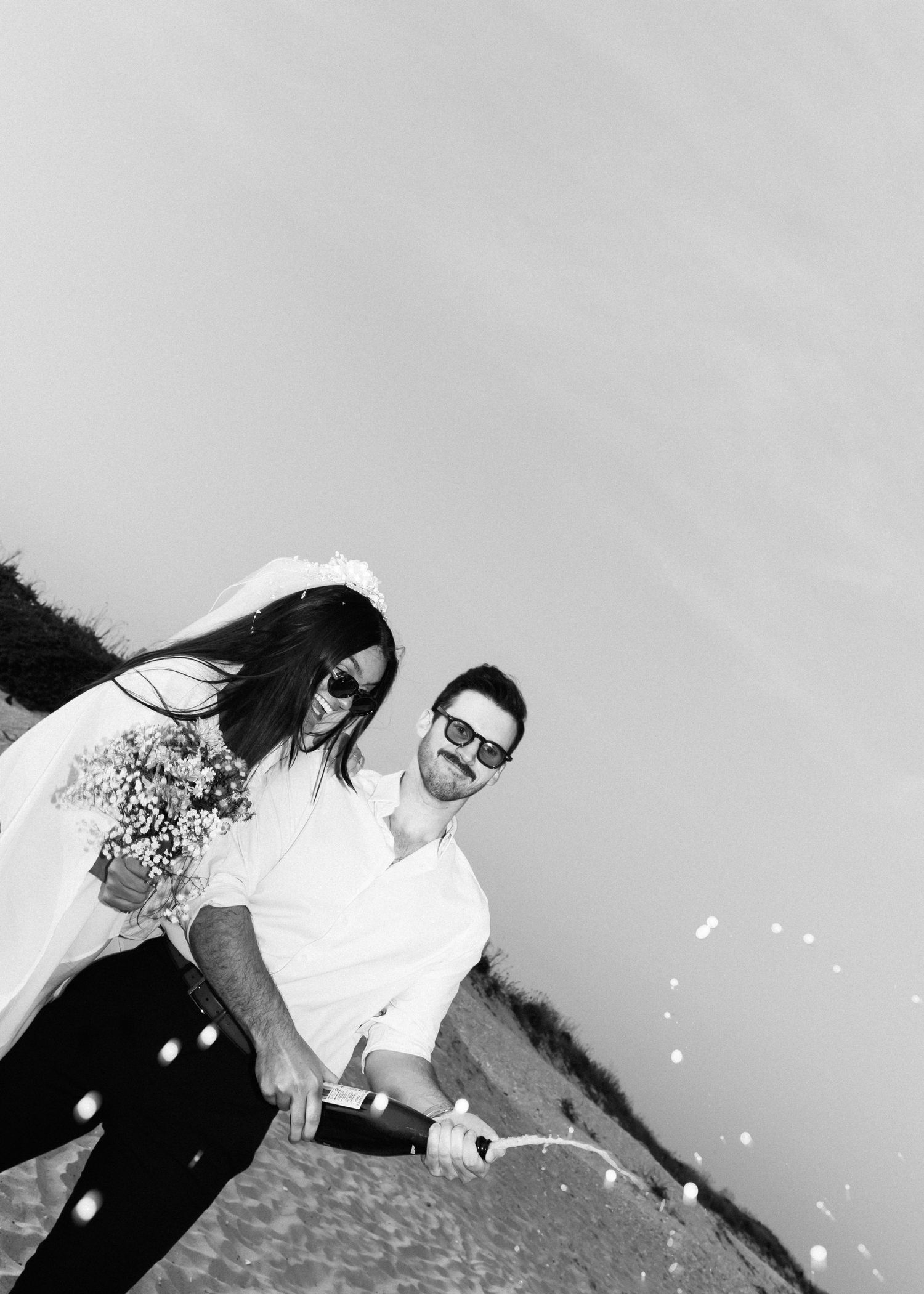A joyful couple in white celebrates by popping champagne on the beach.