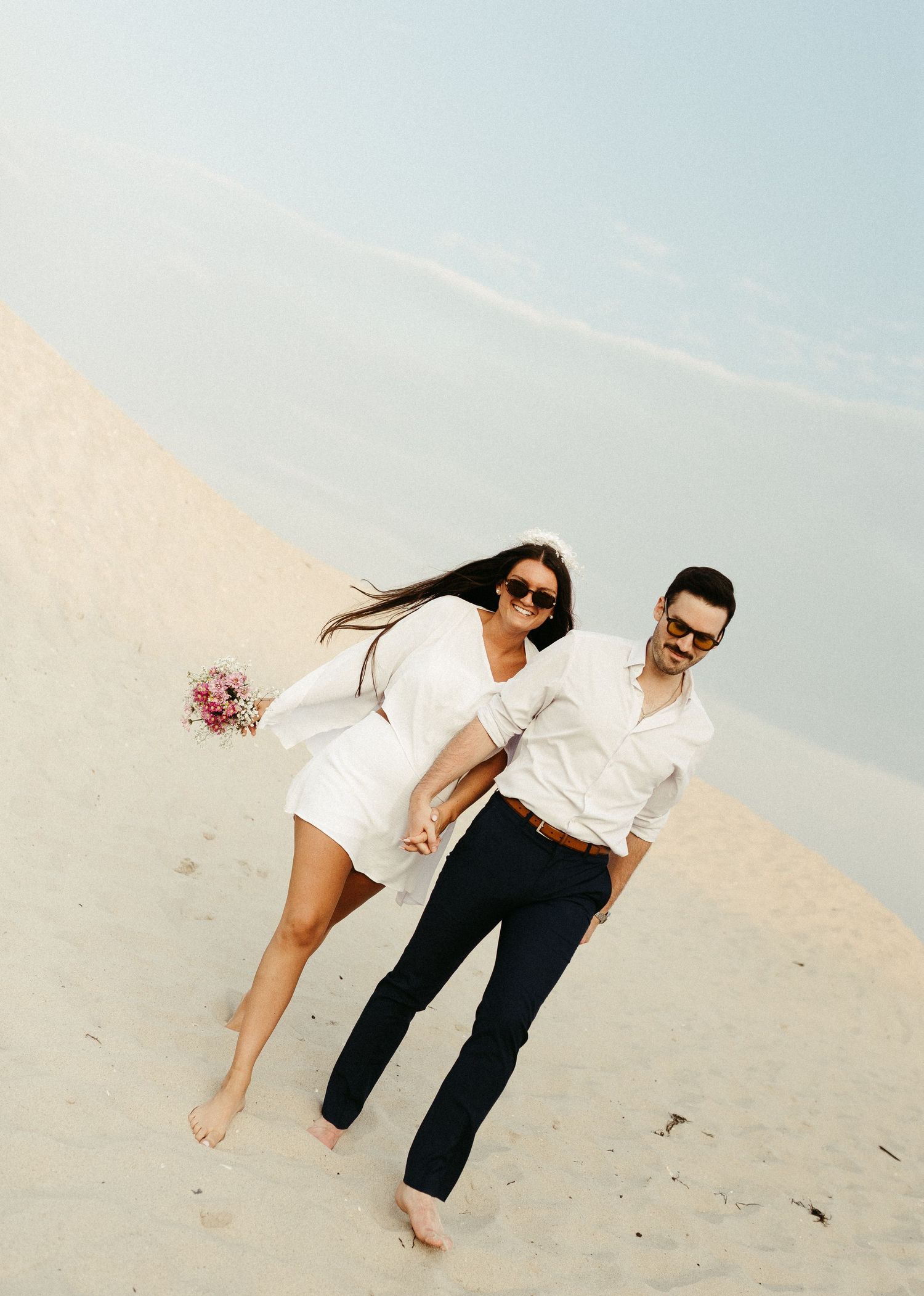 A playful beach wedding photoshoot with a couple in white outfits against sand dunes.
