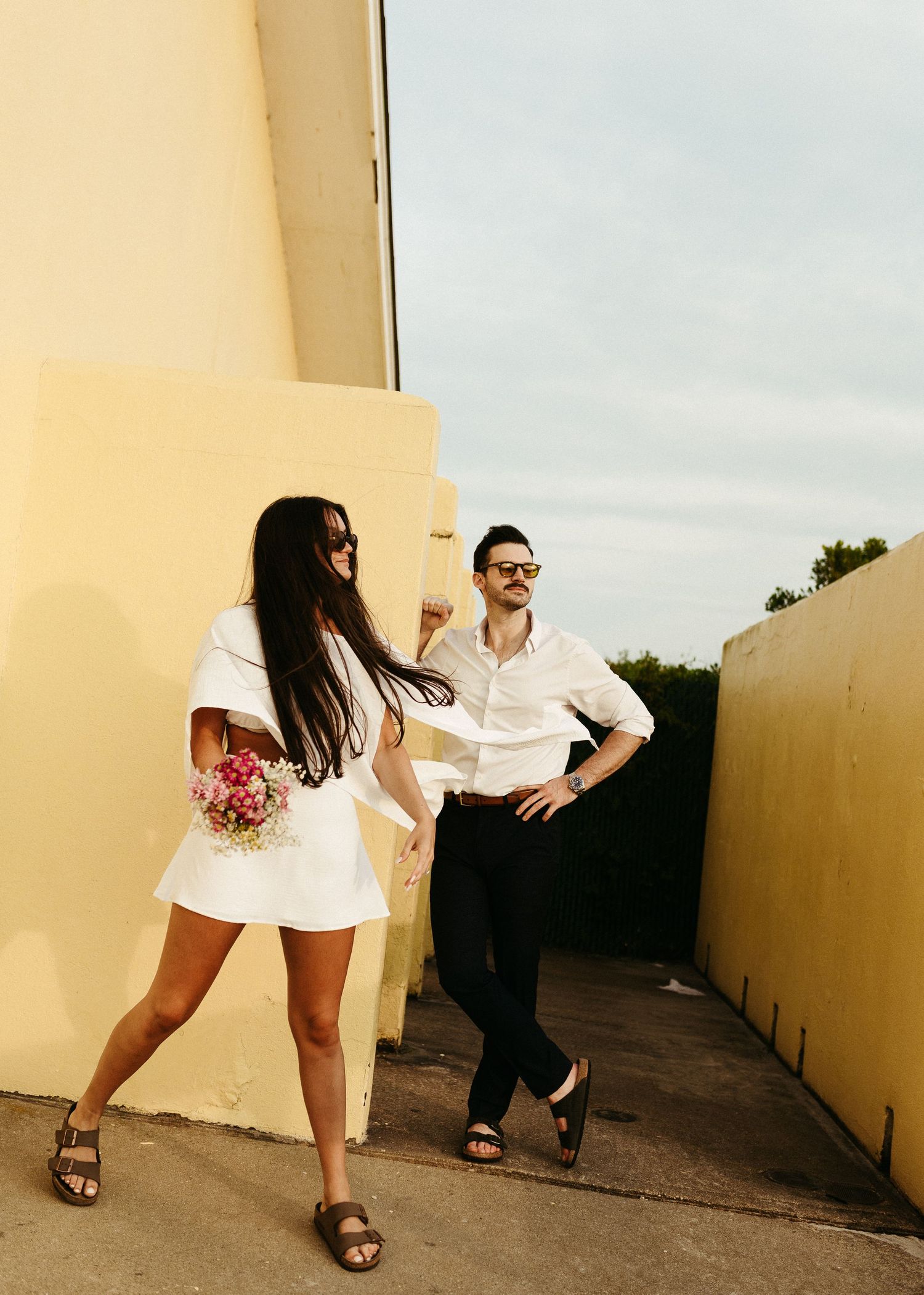 A stylish couple in white outfits poses playfully against a yellow wall.