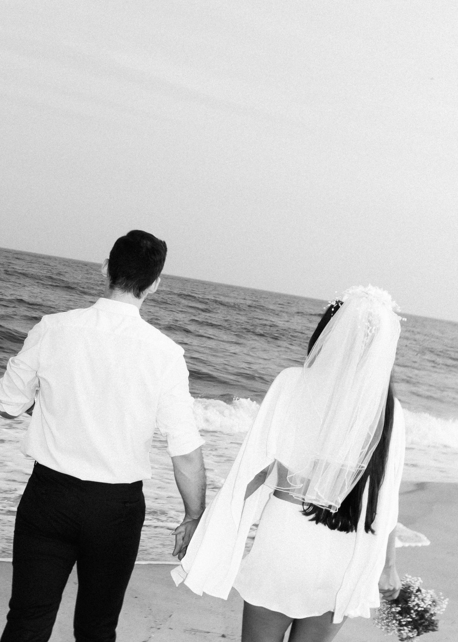 A couple in wedding attire holds hands while walking along a scenic beach at sunset.