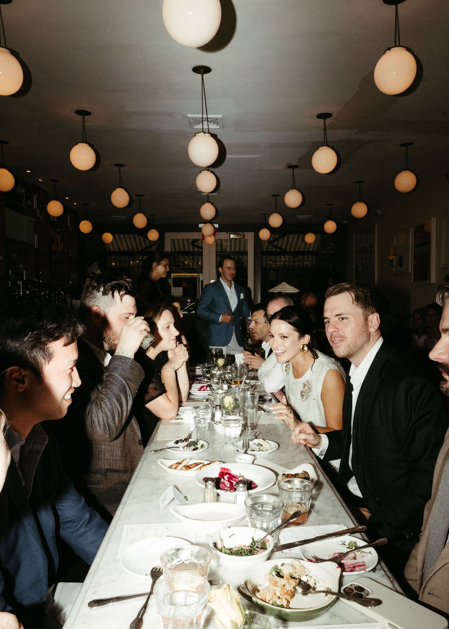Restaurant dining scene with globe lights overhead and guests seated at long table.