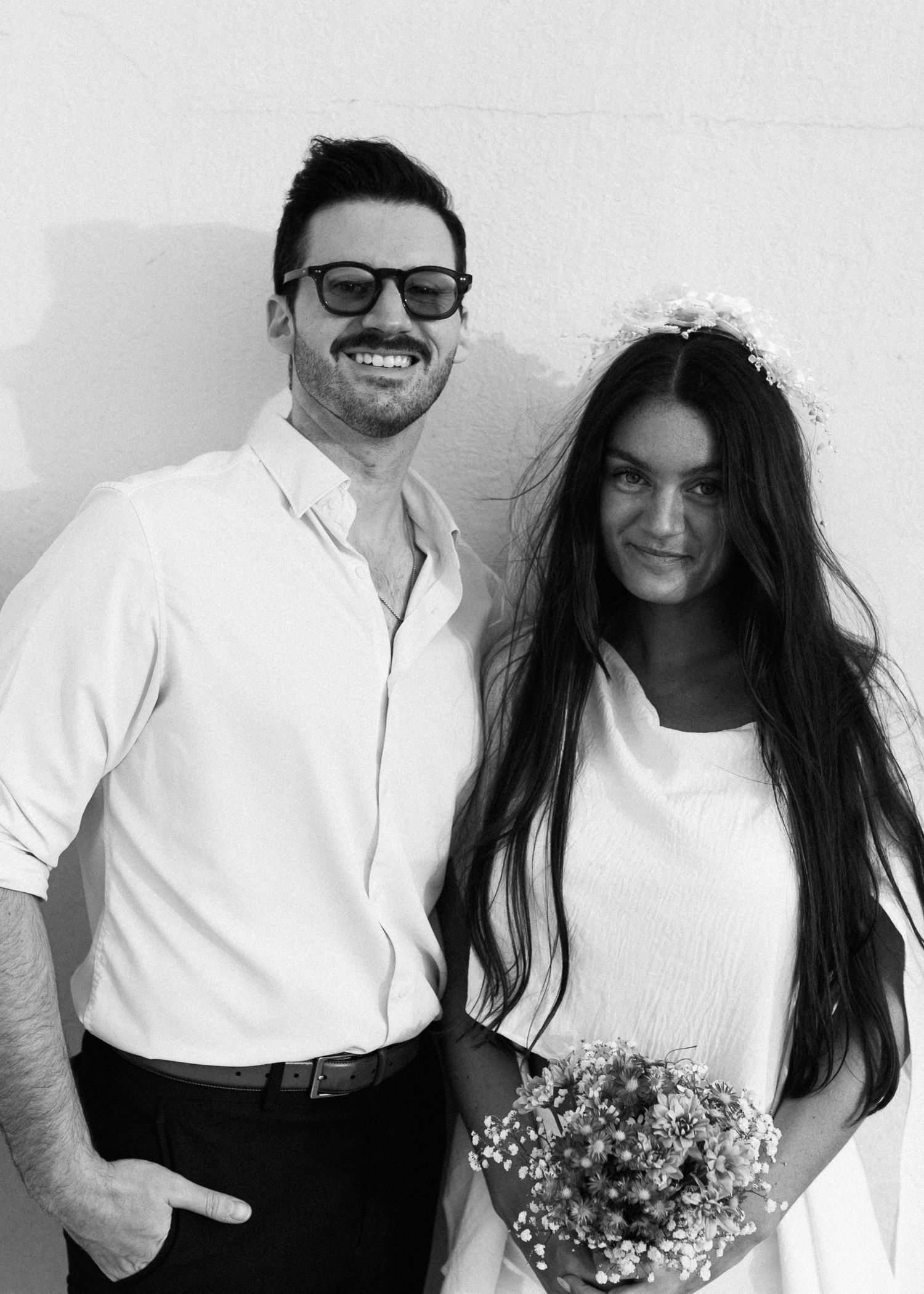 Black and white portrait of couple in matching white outfits with delicate floral bouquet.