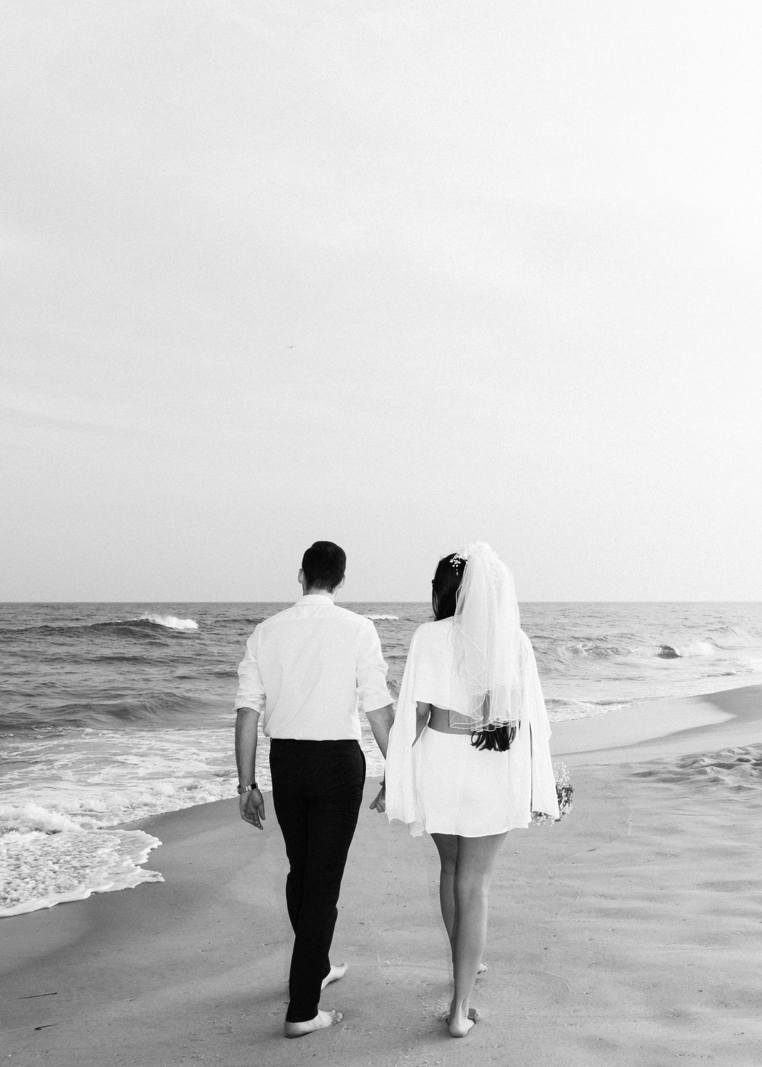 Black and white photo of couple walking barefoot on beach with waves in background.